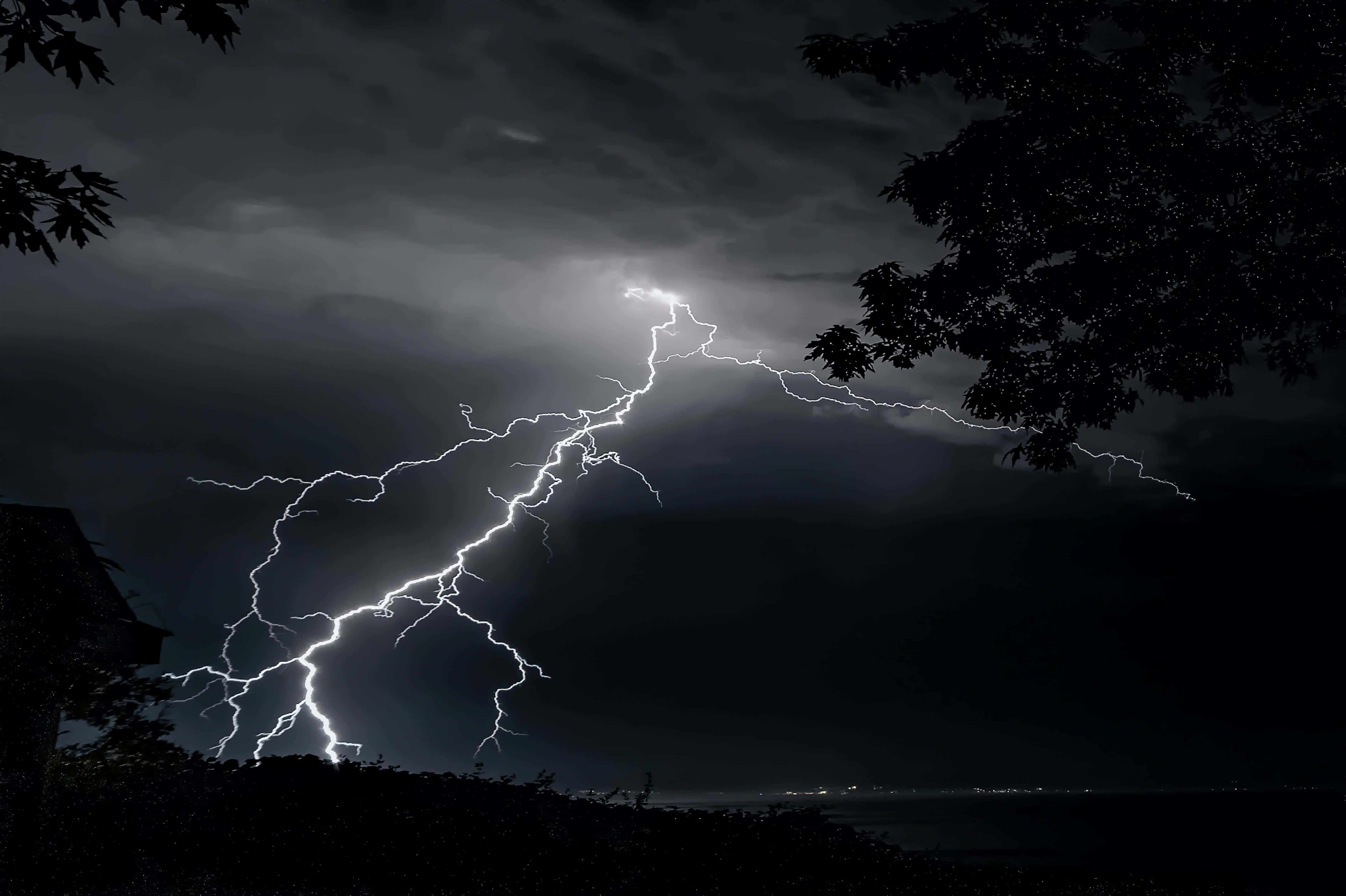 自然的美與力量：暴風雨中的壯觀景象 壁紙 — 一道強勁的閃電在暴風雨的夜空中分叉，襯托著黑暗的樹木剪影和遠處的城市燈光。