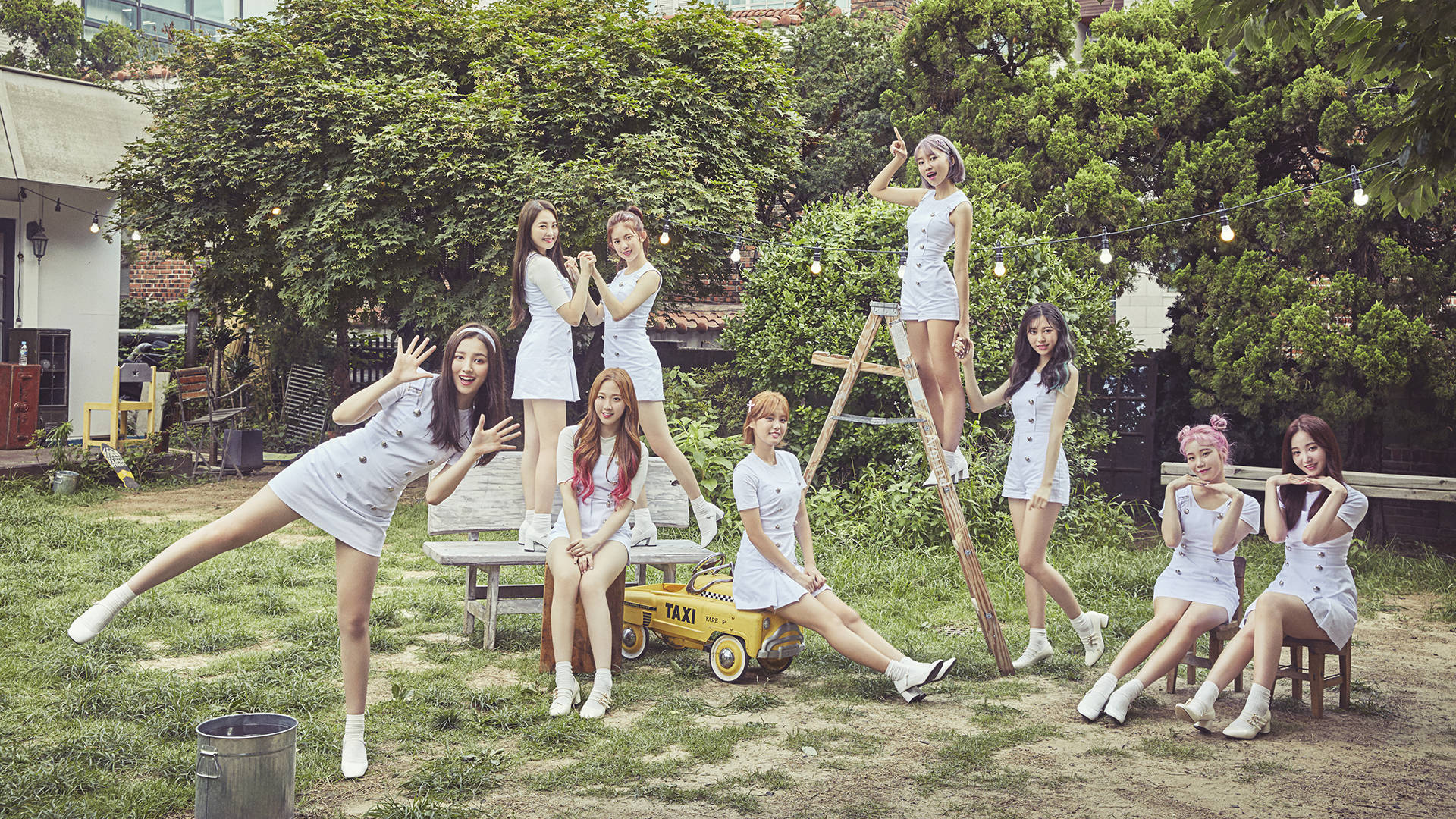 自然隨拍的南西 Momoland 高畫質壁紙 壁紙 — 七位身穿白色服裝的年輕女性在陽光明媚的後院中嬉戲擺姿勢，背景有綠樹、燈串和玩具的士。