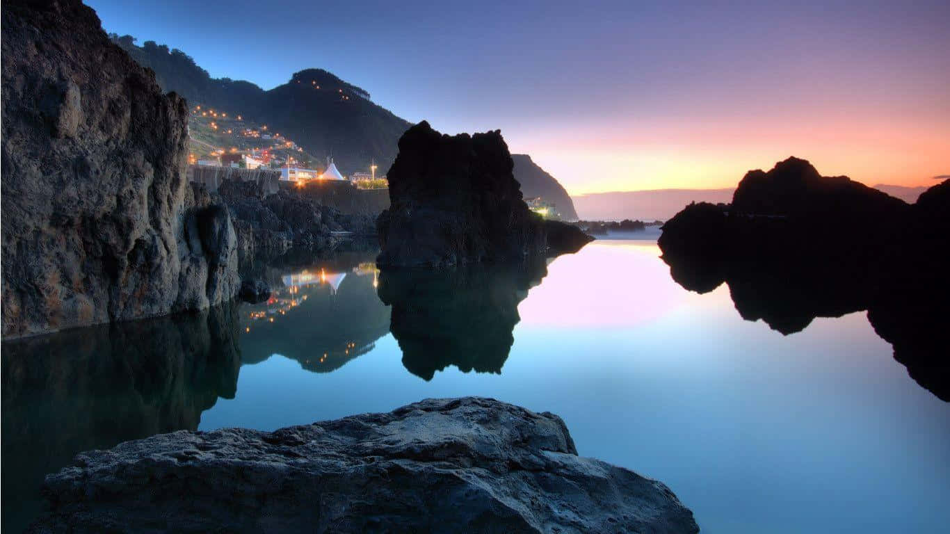 自然風景 1366x768 監視器背景 壁紙 — 寧靜的黃昏場景，村莊燈光閃爍，倒映在平靜的水面上，周圍環繞著深色的火山岩層。
