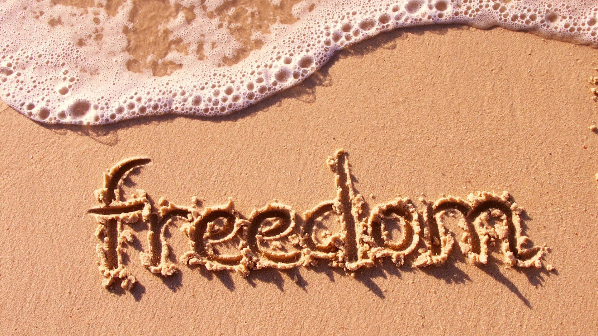 Freedom 桌布 壁紙 — 「自由」這個詞寫在濕沙上，溫柔的海浪帶著白色泡沫正在靠近。