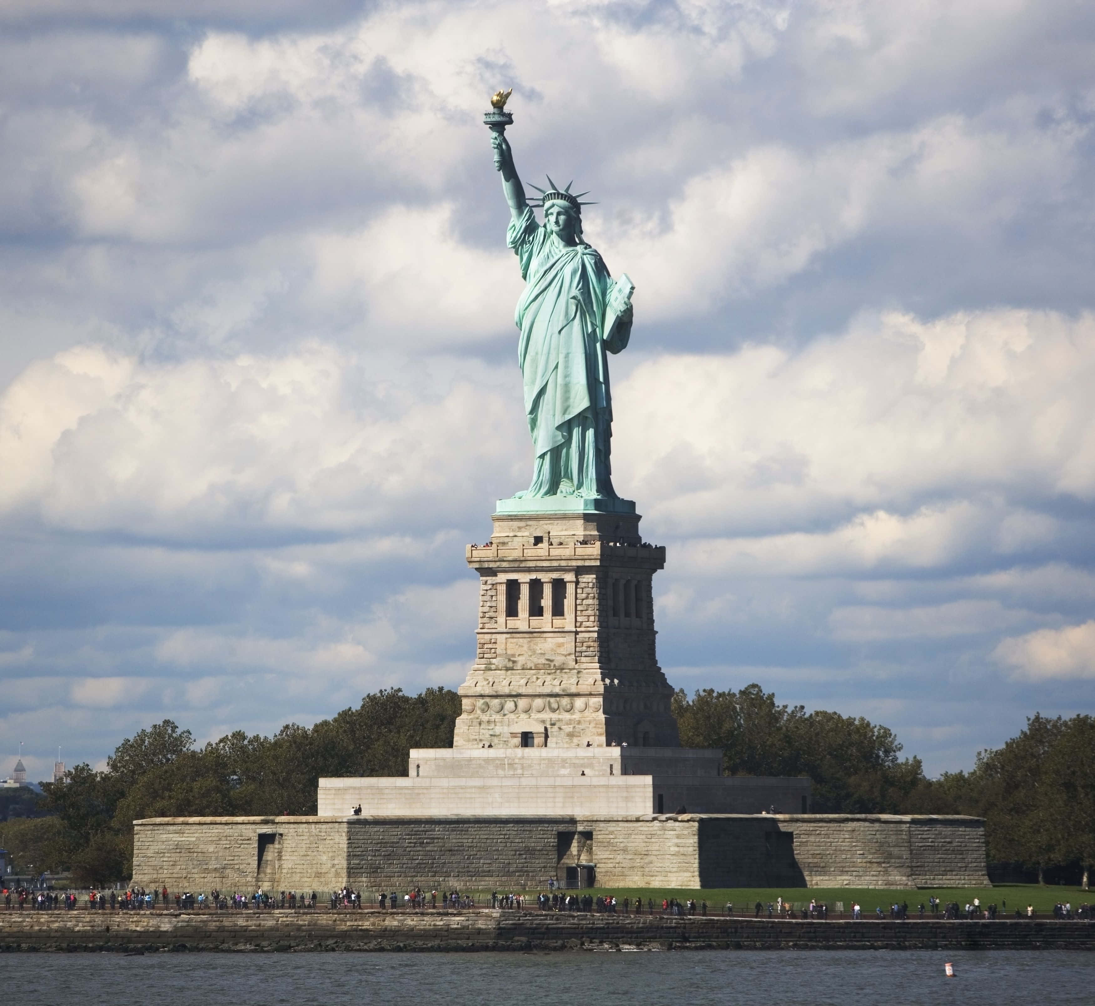 American 桌布 壁紙 — 自由女神像在陰天天空下高高矗立於基座上，人們聚集在水邊。
