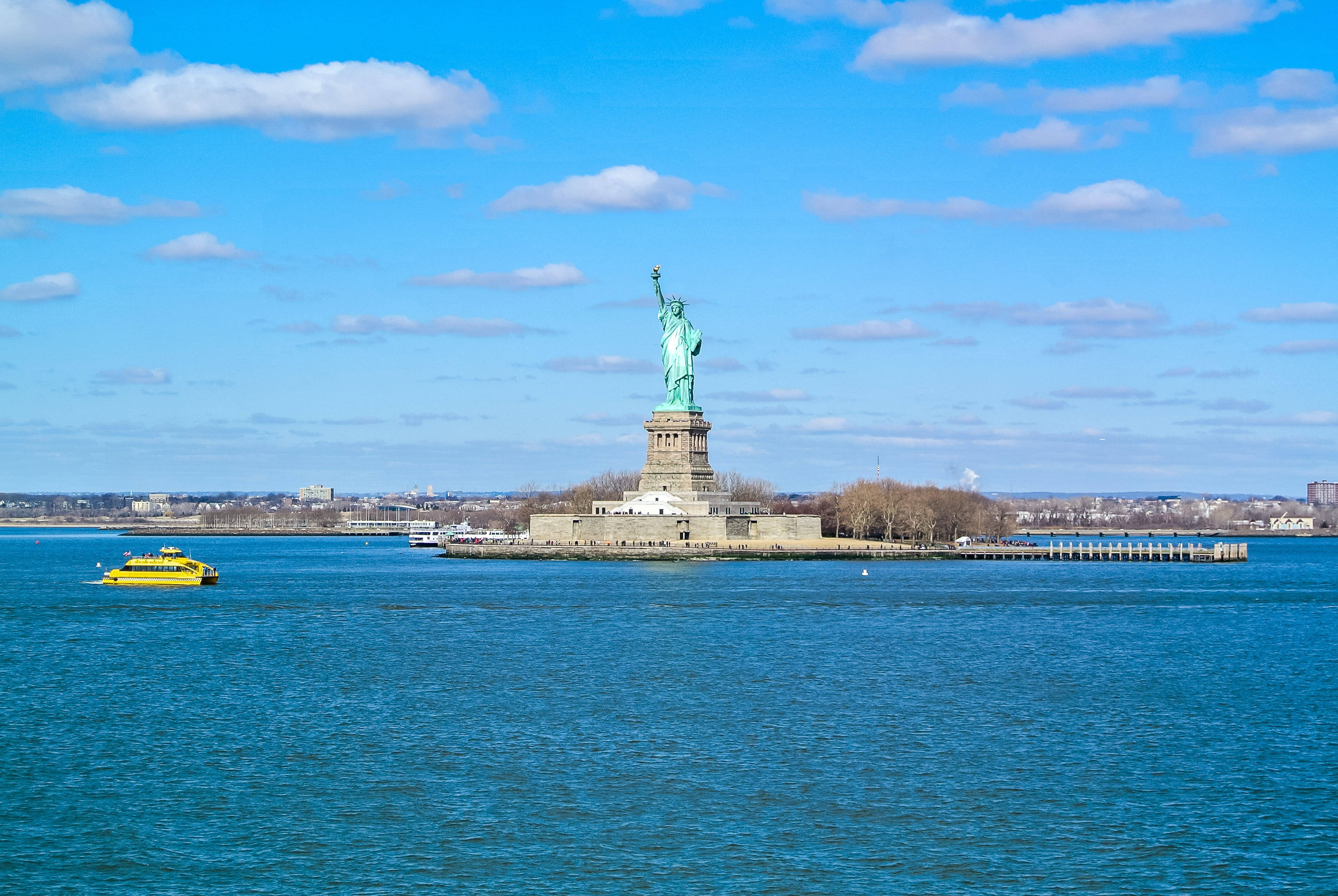 new york 4K 桌布 壁紙 — 自由神像聳立在自由島上，天空湛藍，白雲點綴，一艘黃色渡輪在深藍色的水面上滑行。