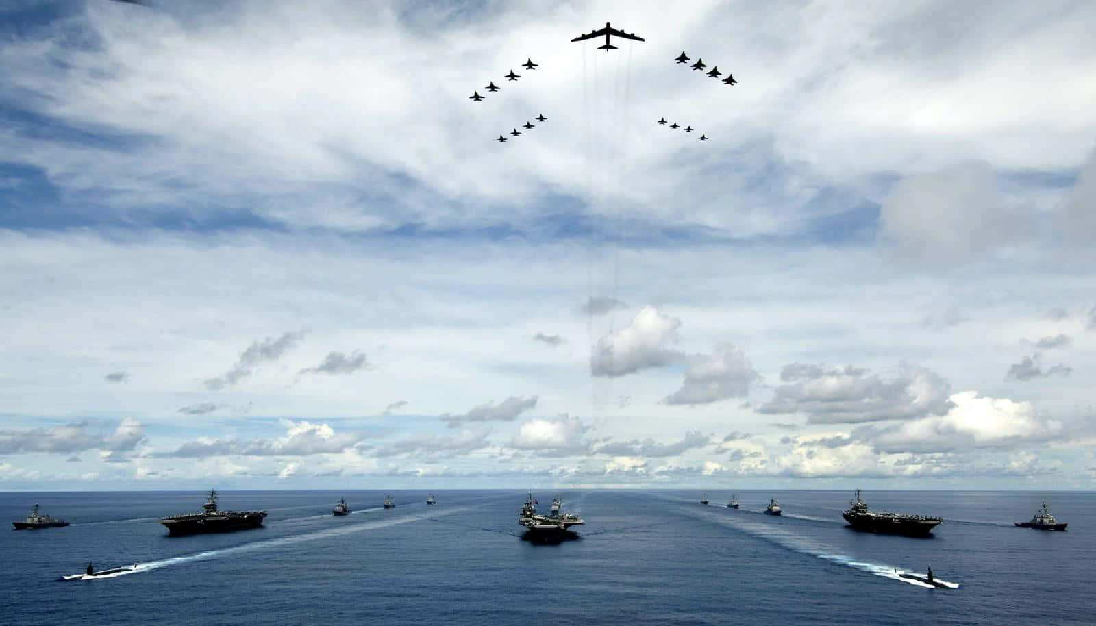 自豪的海軍退伍軍人向美國國旗敬禮 壁紙 — 一支由航空母艦和軍艦組成的海軍艦隊，在陰天之下，戰鬥機在上空飛過，編隊航行在廣闊的藍色海洋上。