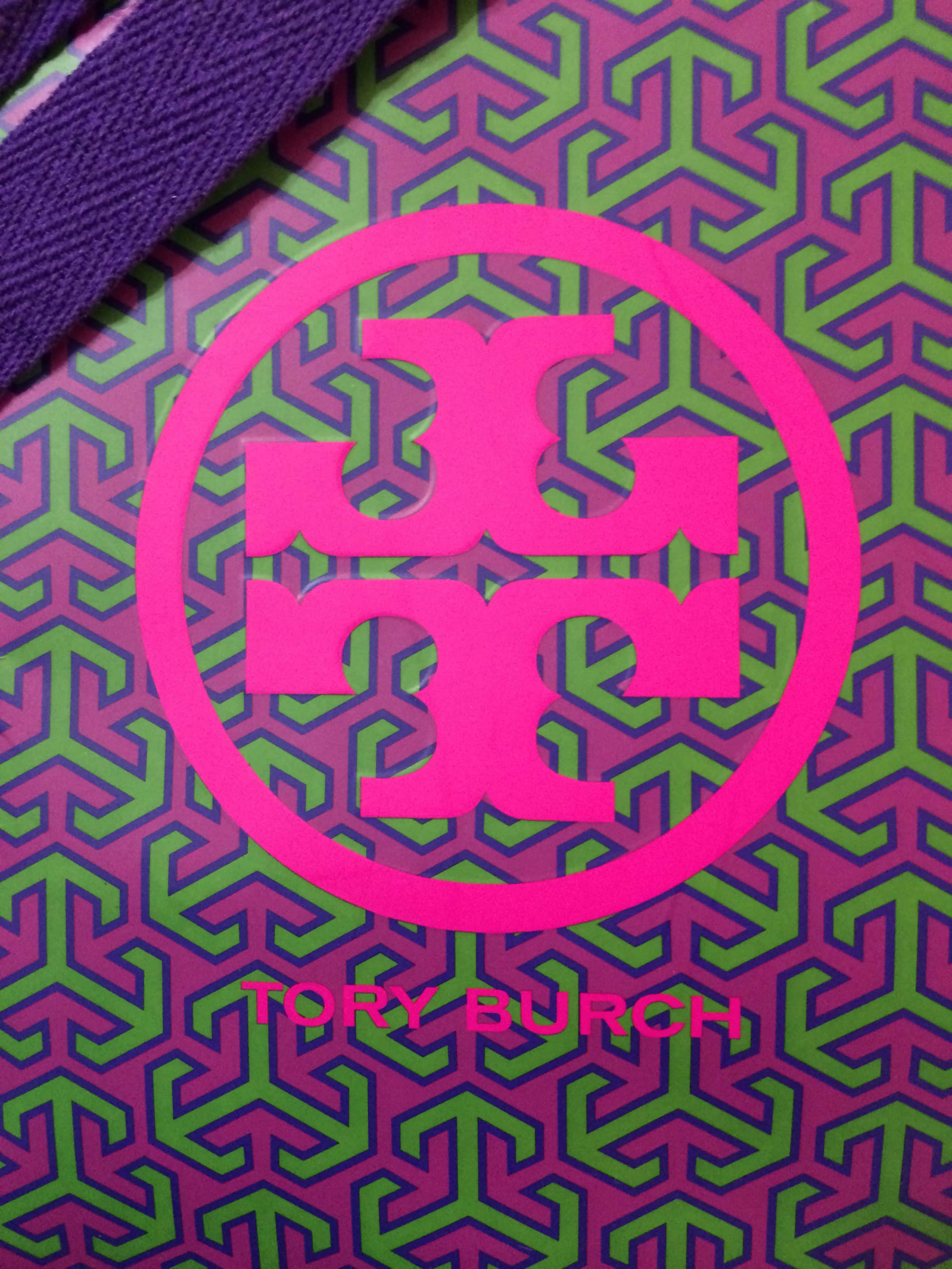 致敬 Tory Burch 的經典時尚與品味 壁紙 — 紫色和綠色幾何圖案手袋上鮮豔粉紅色Tory Burch標誌的特寫。