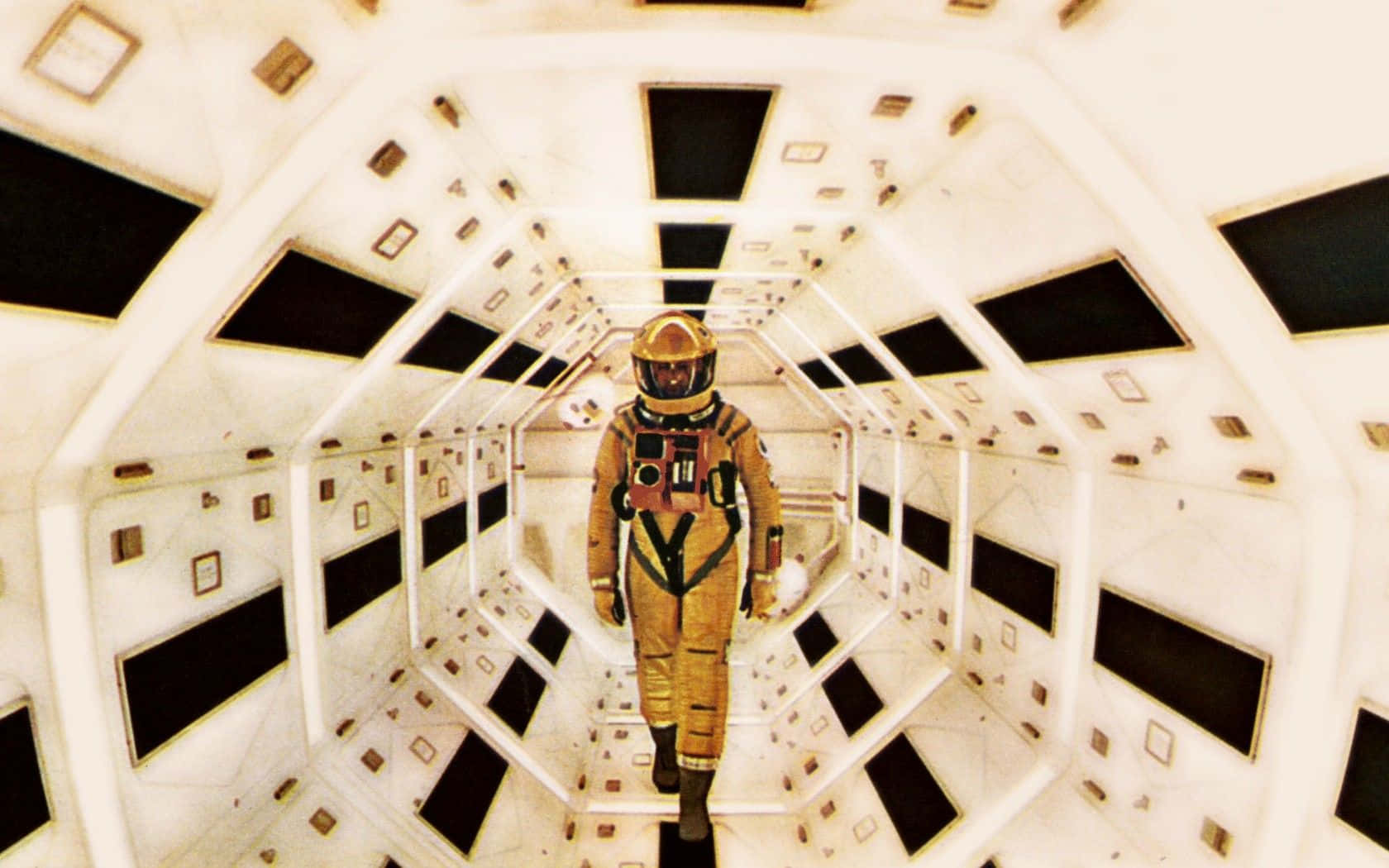 與《2001太空漫遊》一同超越想像的旅程 壁紙 — 一名身穿金色太空衣的太空人，沿著一條狹窄的白色六角形太空船走廊行走，走廊上有黑色面板和小型方形裝置。