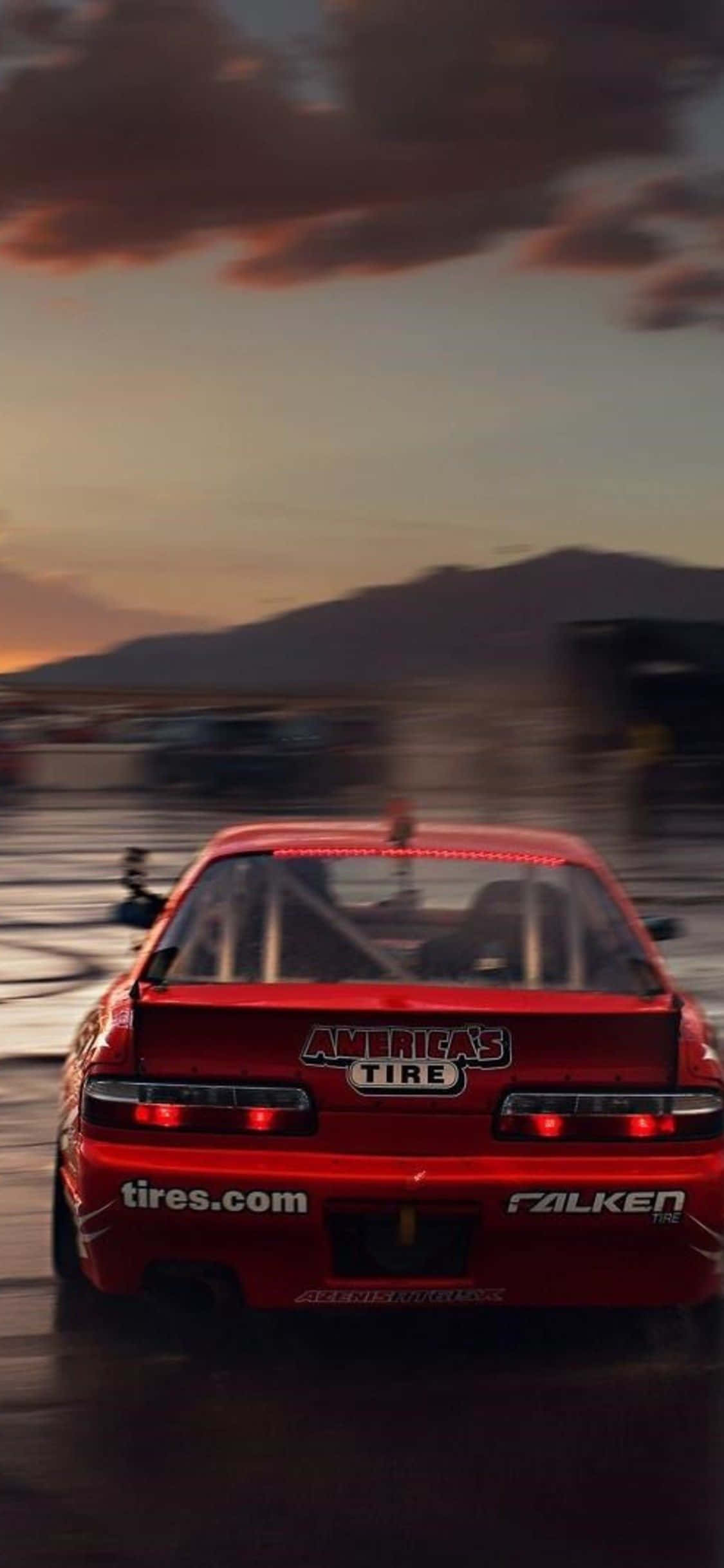 與 iPhone Xs Max 一起體驗《Project Cars 2》的刺激 壁紙 — 一輛紅色賽車在日落時分的潮濕賽道上疾馳而去，背景有山巒，呈現動態模糊效果。