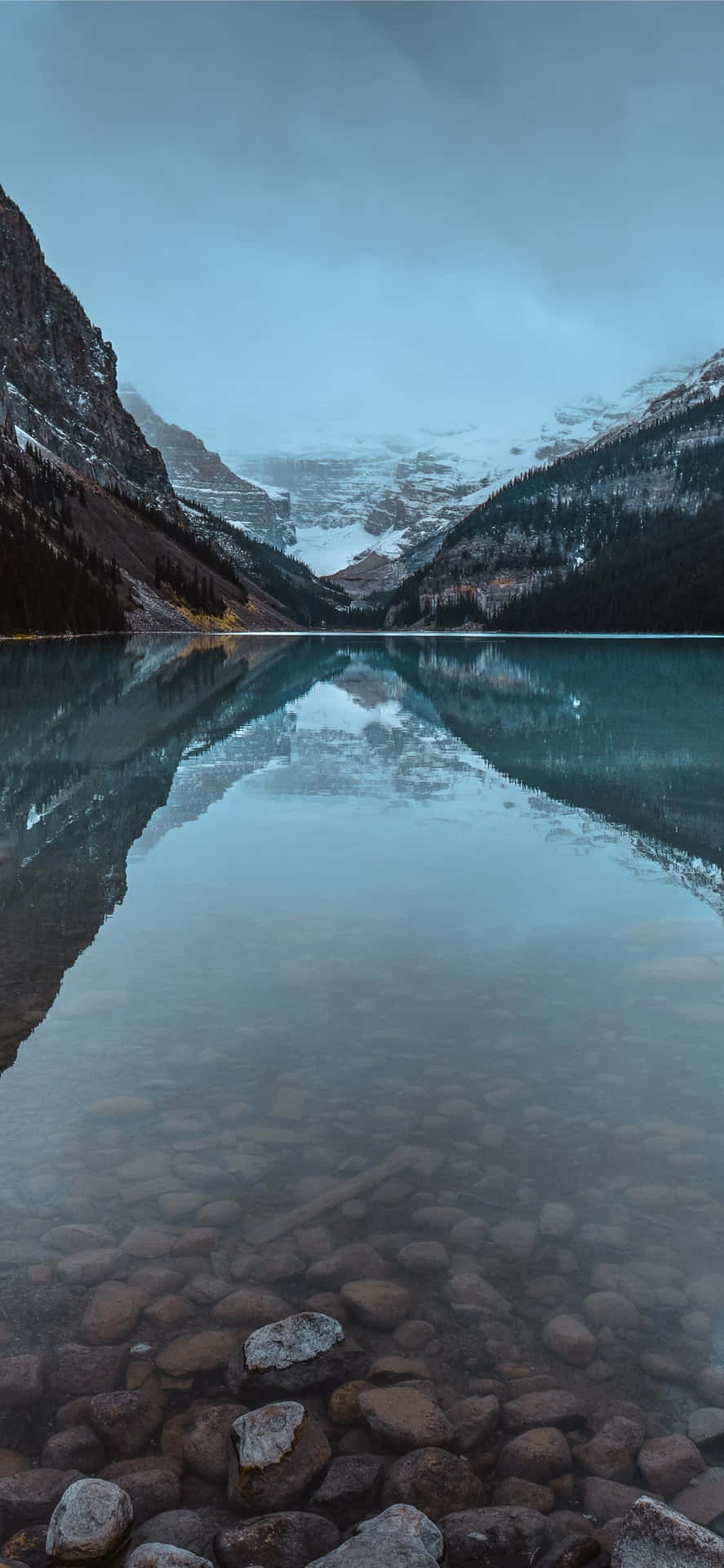 山湖在多雲的天空下映照積雪的山峰，淺水中可見岩石。
