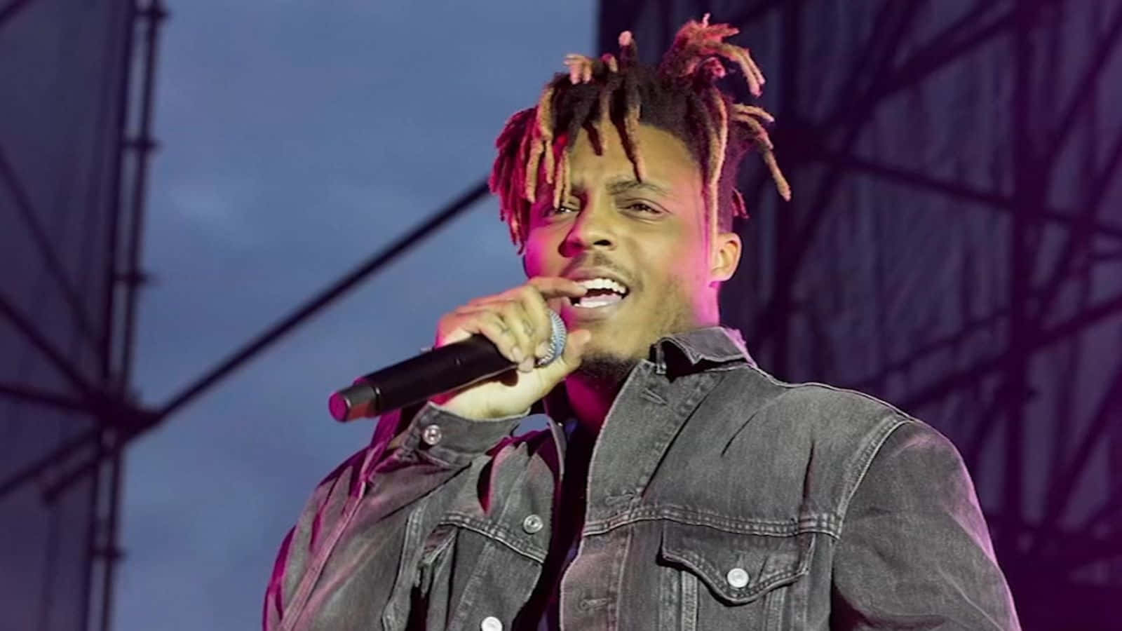 與 Juice Wrld 一同享受嘻哈與現場音樂 壁紙 — 一位擁有彩色辮子的男歌手在舞台燈光下熱情地對著麥克風表演。