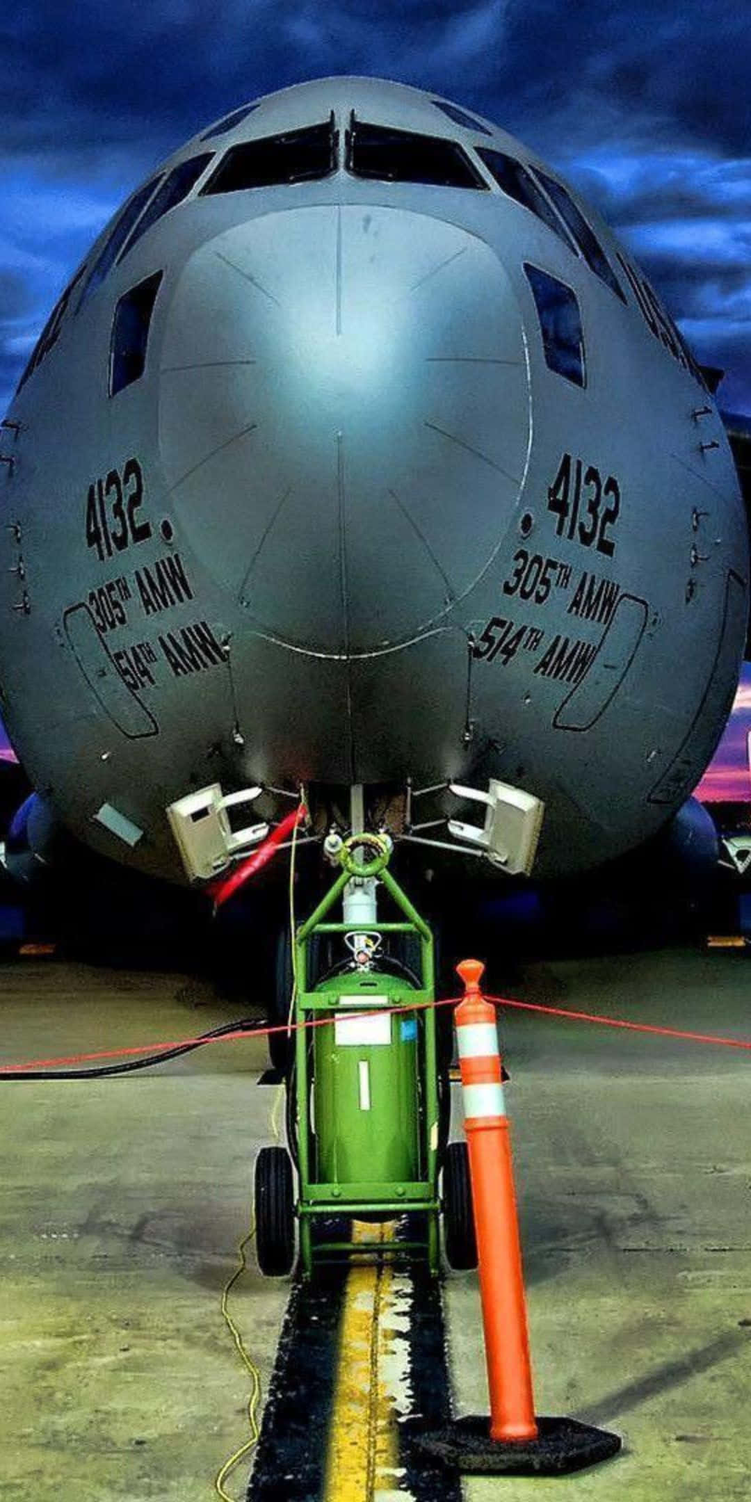 與 Pixel 3 一同穿越雲霄 壁紙 — 灰色軍用運輸機機頭近距離特寫，帶有維修設備和橙色錐體，在戲劇性的暮色天空下的停機坪上。