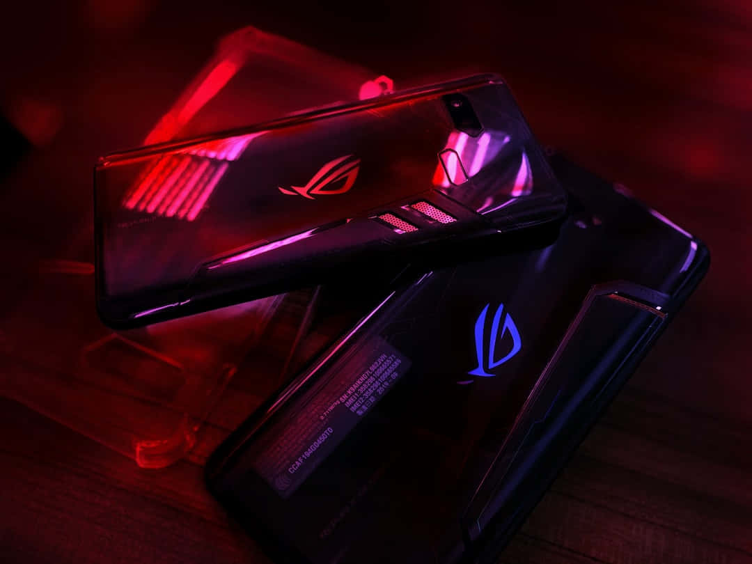與 ROG 一起投入遊戲 壁紙 — 兩部帶有發光RGB燈的遊戲手機放在木質表面上，在戲劇性的紅色和紫色燈光下。