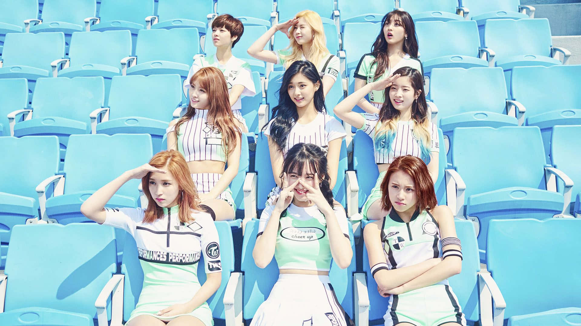與 Twice 一起感受特別的感覺 壁紙 — 九位年輕女性穿著相配的運動服，在明亮的藍色體育場座位上，在晴朗的日光下俏皮地擺姿勢。
