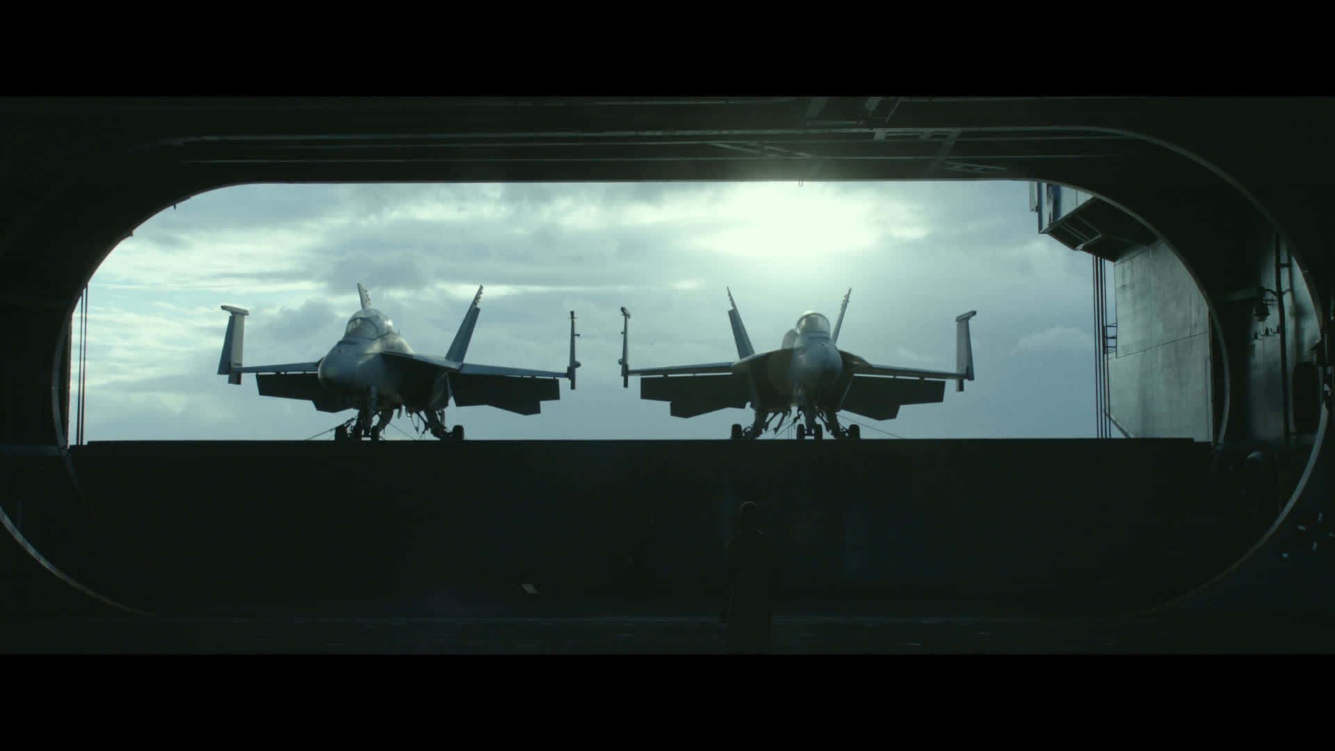 與《頂尖飛行員》一同翱翔天際 壁紙 — 兩架戰鬥機在航空母艦甲板上以剪影形式站立，透過彎曲的舷窗觀看，天空多雲。