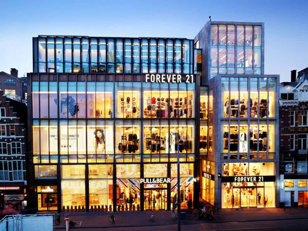 與Forever 21一起迎接美麗時尚季節 壁紙 — 一間現代玻璃幕牆的Forever 21和Pull&Bear商店在黃昏時分溫暖地發光，映襯著暮色天空。