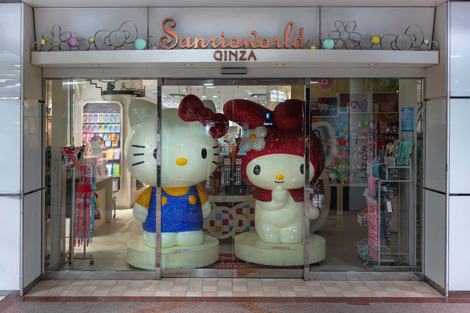 與Hello Kitty共度陽光明媚的下午 壁紙 — 兩個巨大的閃亮Hello Kitty雕像矗立在Sanrio World Ginza的玻璃入口處，周圍環繞著色彩繽紛的商品。