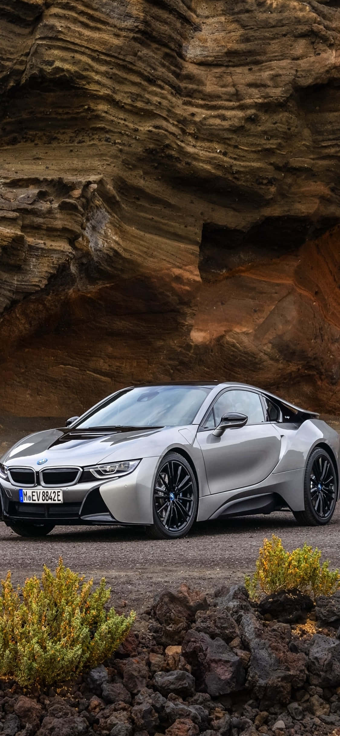 與iPhone X與BMW一同享受奢華駕馭 壁紙 — 一輛時尚的銀色BMW i8跑車停泊在戲劇性的層狀岩層下，周圍有稀疏的綠色灌木。