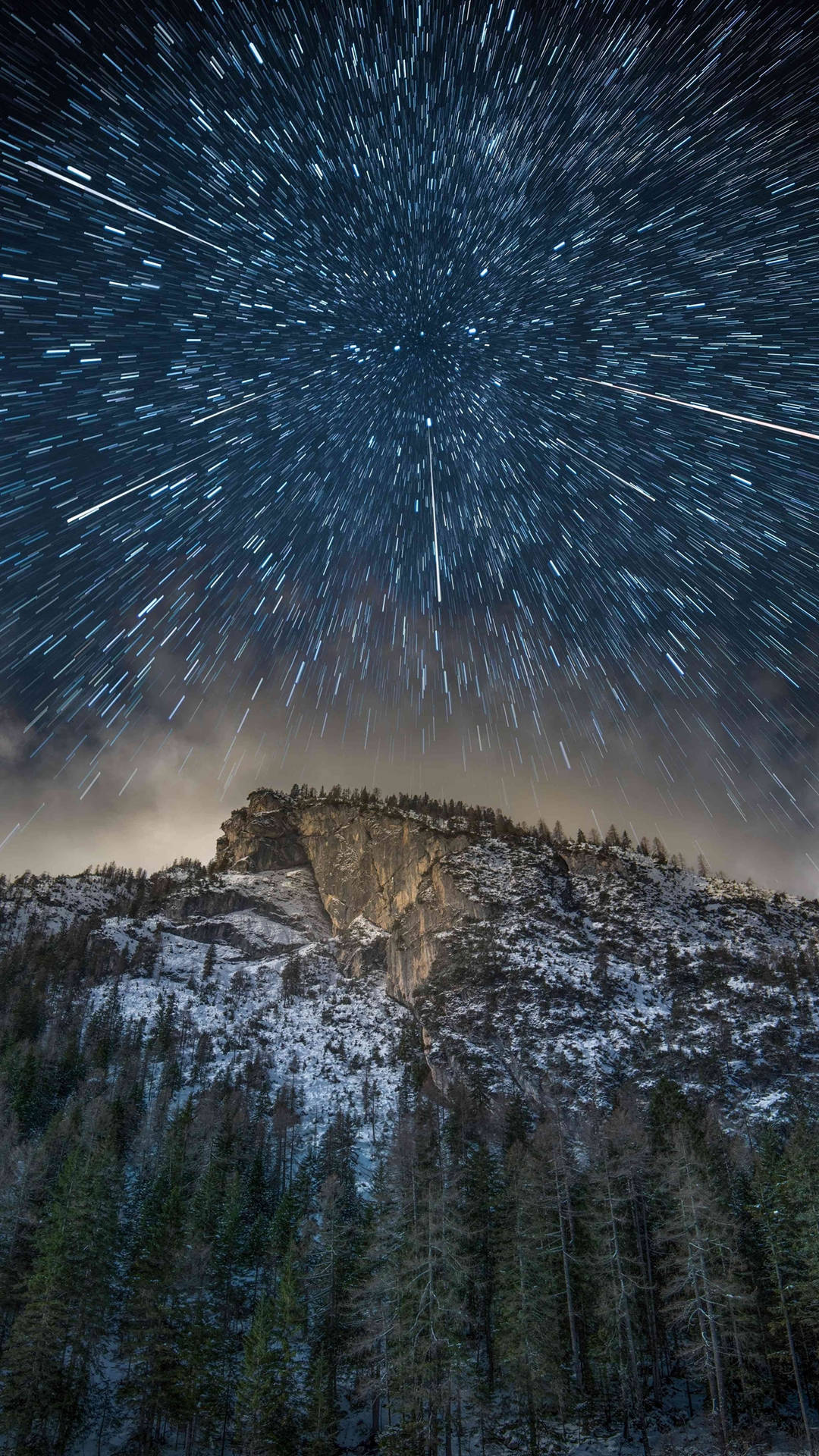 夜間白雪覆蓋的山峰和松樹林上方的星軌。