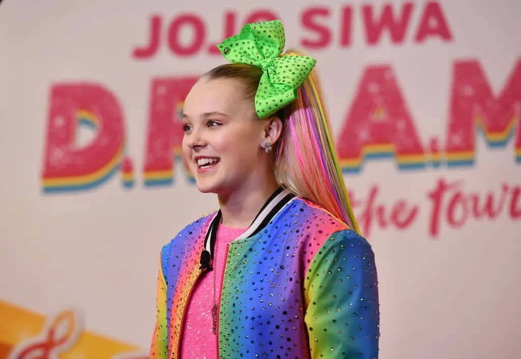 與Jojo一同欣賞世界展開 壁紙 — JoJo Siwa 穿著彩虹閃片外套，色彩繽紛的馬尾上繫著一個巨大的綠色波點蝴蝶結，露出燦爛的笑容。