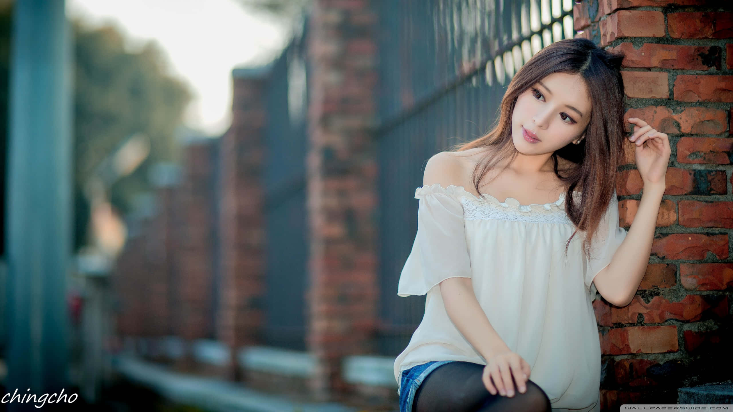 與可愛女孩共度歡樂桌面時光 壁紙 — 一位年輕女性身穿白色露肩襯衫，靠在一面風化的磚牆上，柔和陽光穿過她身後的樹木灑落。