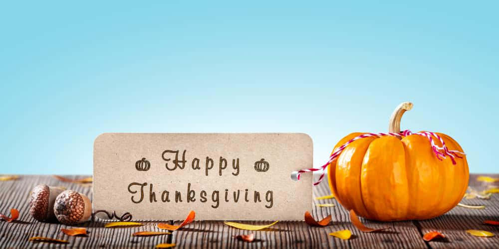 與家人共慶感恩節並祈禱祝福 壁紙 — 一個質樸的「Happy Thanksgiving」標籤放在一個鮮橙色南瓜和橡子旁邊，木質表面上散落著秋葉，背景是清澈的藍天。