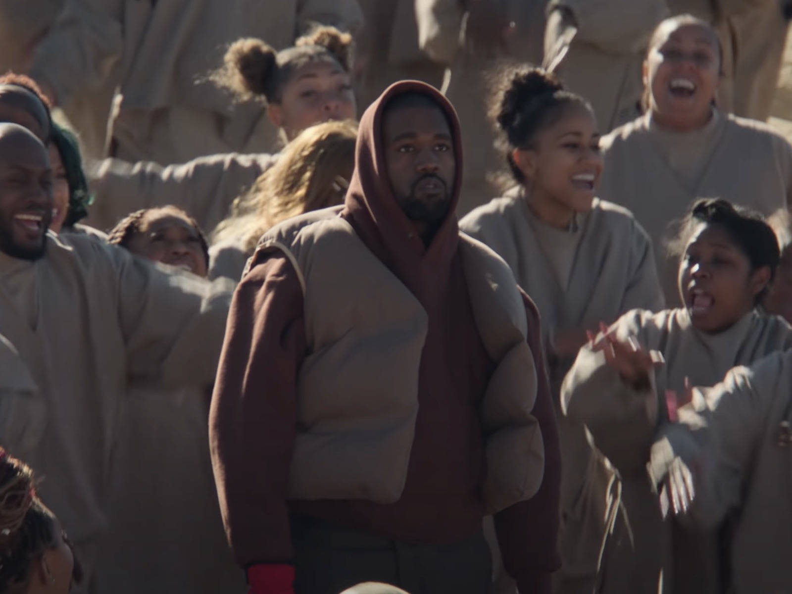 與已故母親Donda共度難忘時刻的坎耶·威斯特 壁紙 — Kanye West 身穿栗色連帽衫，在溫暖的陽光下，莊嚴地站在一群穿著米色長袍的歡樂人群中。