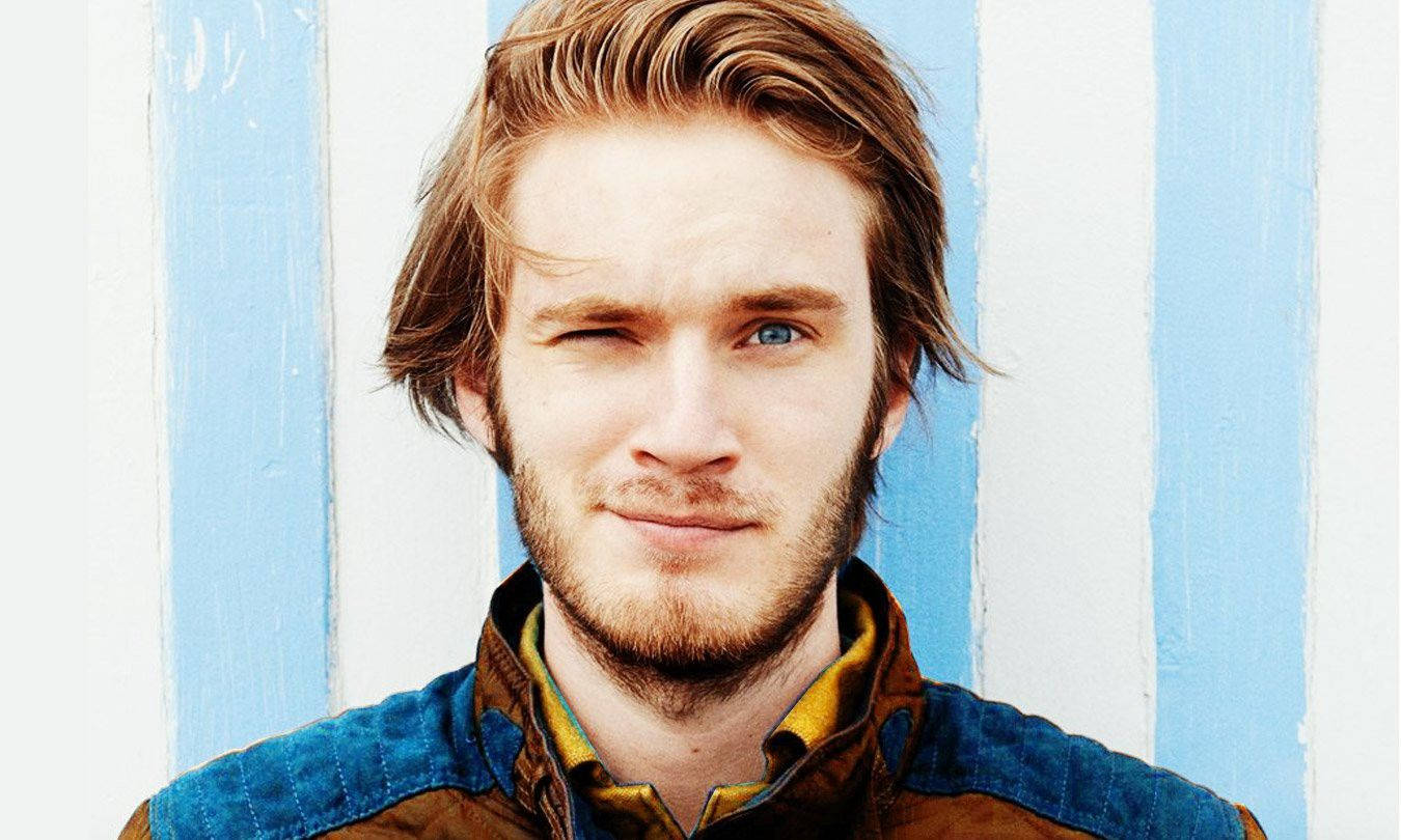 「與我一同踏上 Sub2PewDiePie 旅程！」 壁紙 — 一位擁有引人注目的藍色眼睛和微紅頭髮的年輕男子，在藍白條紋牆前頑皮地眨眼。