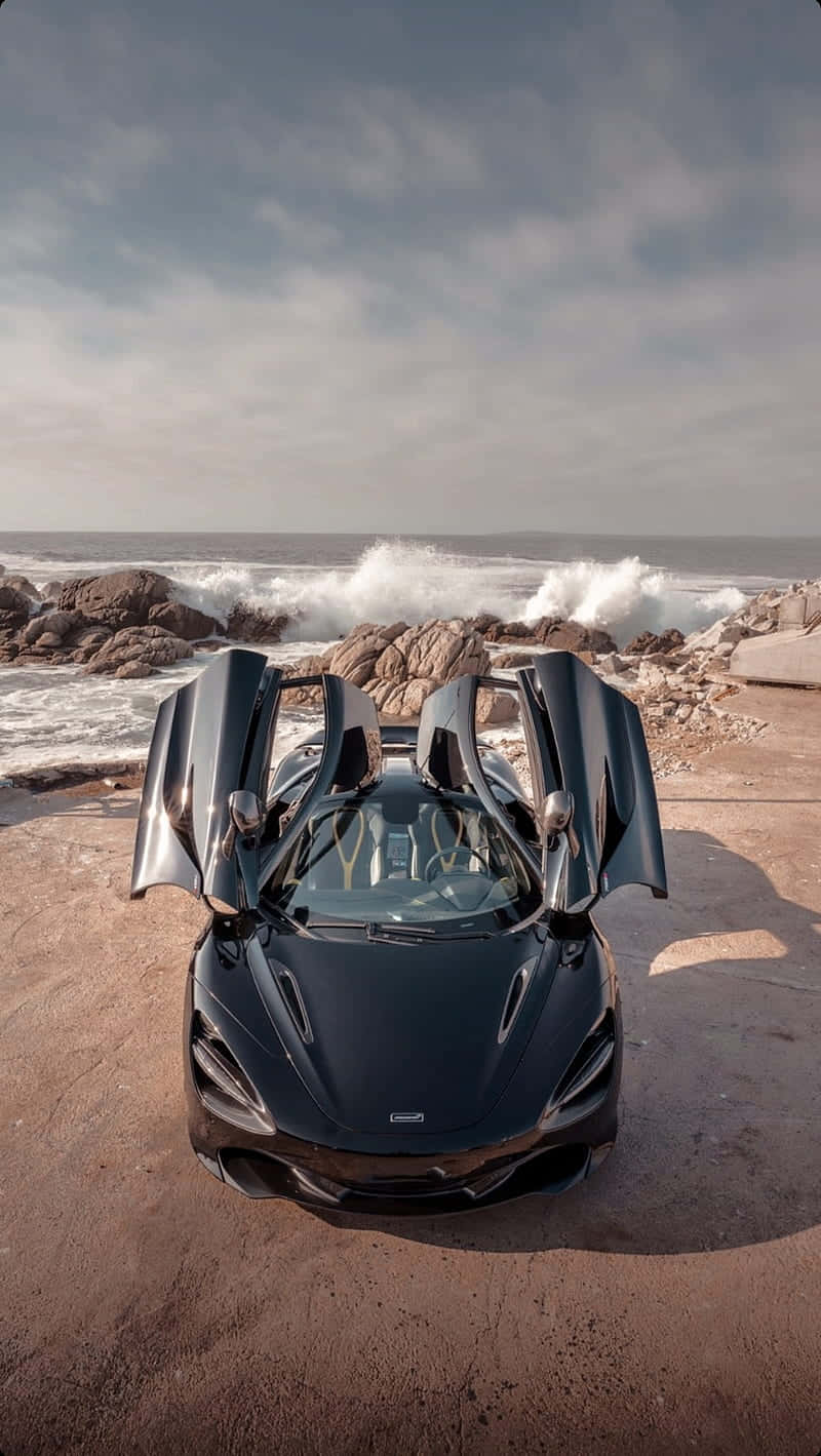 與新款 iPhone Xs Max 和《Project Cars 2》在賽道上虛擬競速 壁紙 — 一架流線型黑色跑車，蝴蝶門敞開，停泊在崎嶇的海岸懸崖上，俯瞰著洶湧的海浪。