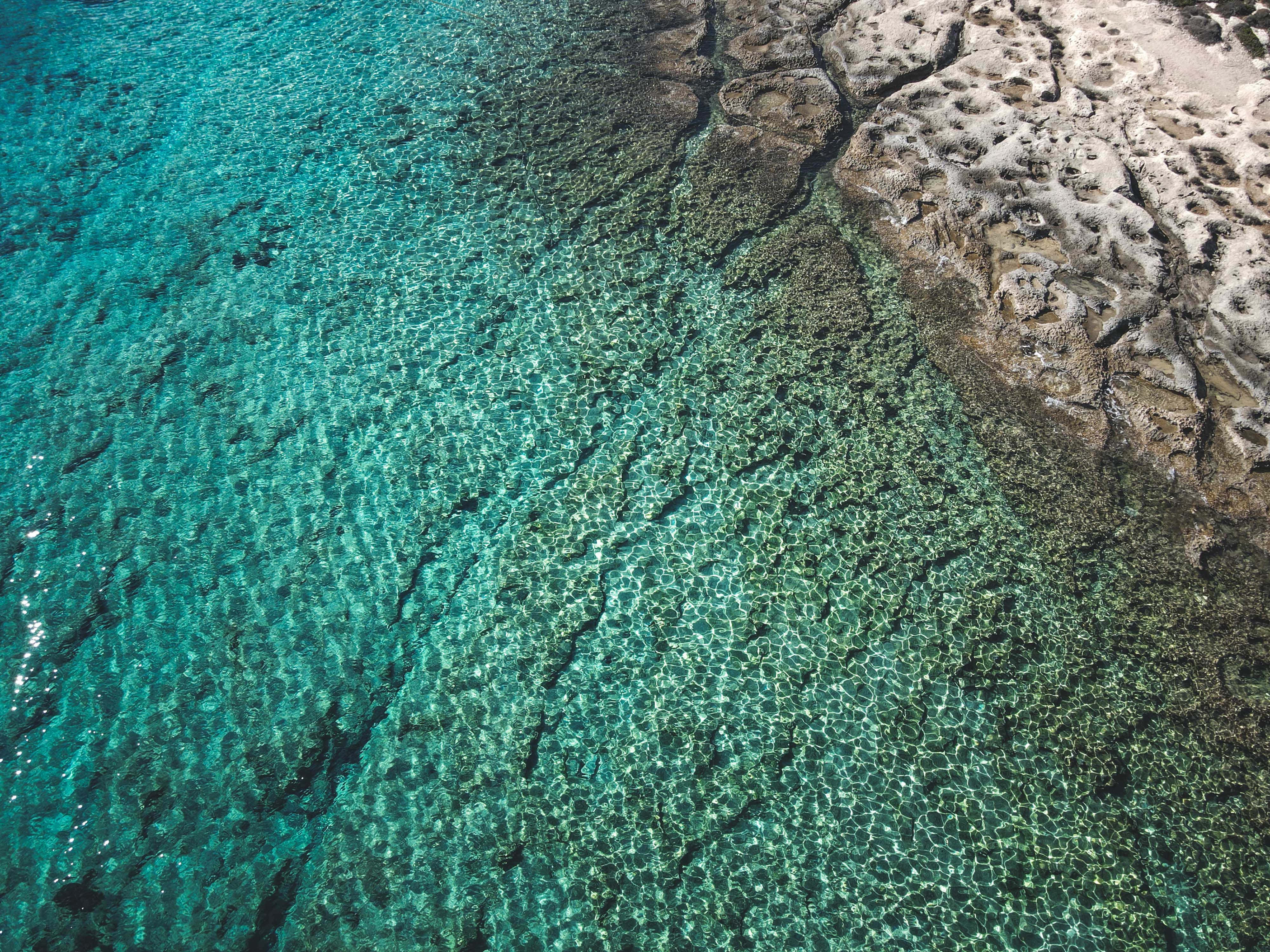 與朋友共賞海灘美景 壁紙 — 從空中俯瞰清澈的碧綠海水，露出鮮豔的珊瑚礁圖案，旁邊是崎嶇的、被陽光曬白的岩石 formations。
