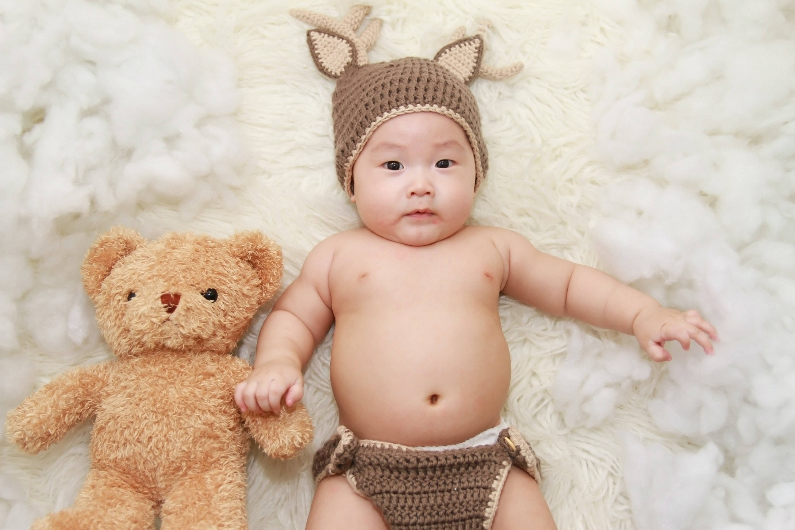 與泰迪熊相伴的可愛嬰兒 壁紙 — 一個戴著棕色鉤針編織鹿帽的嬰兒躺在柔軟的白色地毯上，旁邊有一隻相配的泰迪熊。