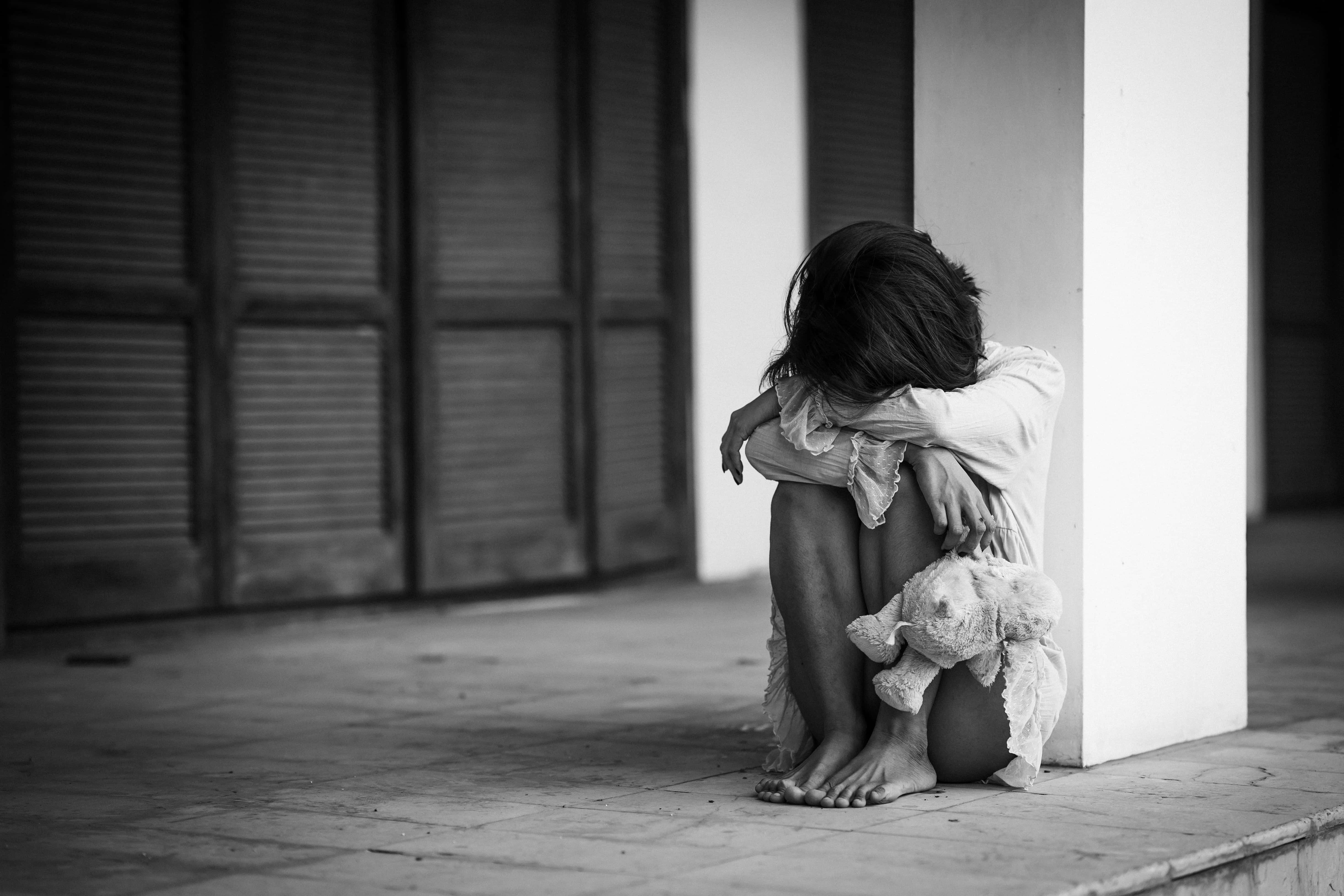 與泰迪熊相伴的悲傷女子照片 壁紙 — 一個孩子蜷縮在瓷磚地板上靠著柱子，緊抓著一隻破舊的泰迪熊，畫面呈鮮明的黑白對比。