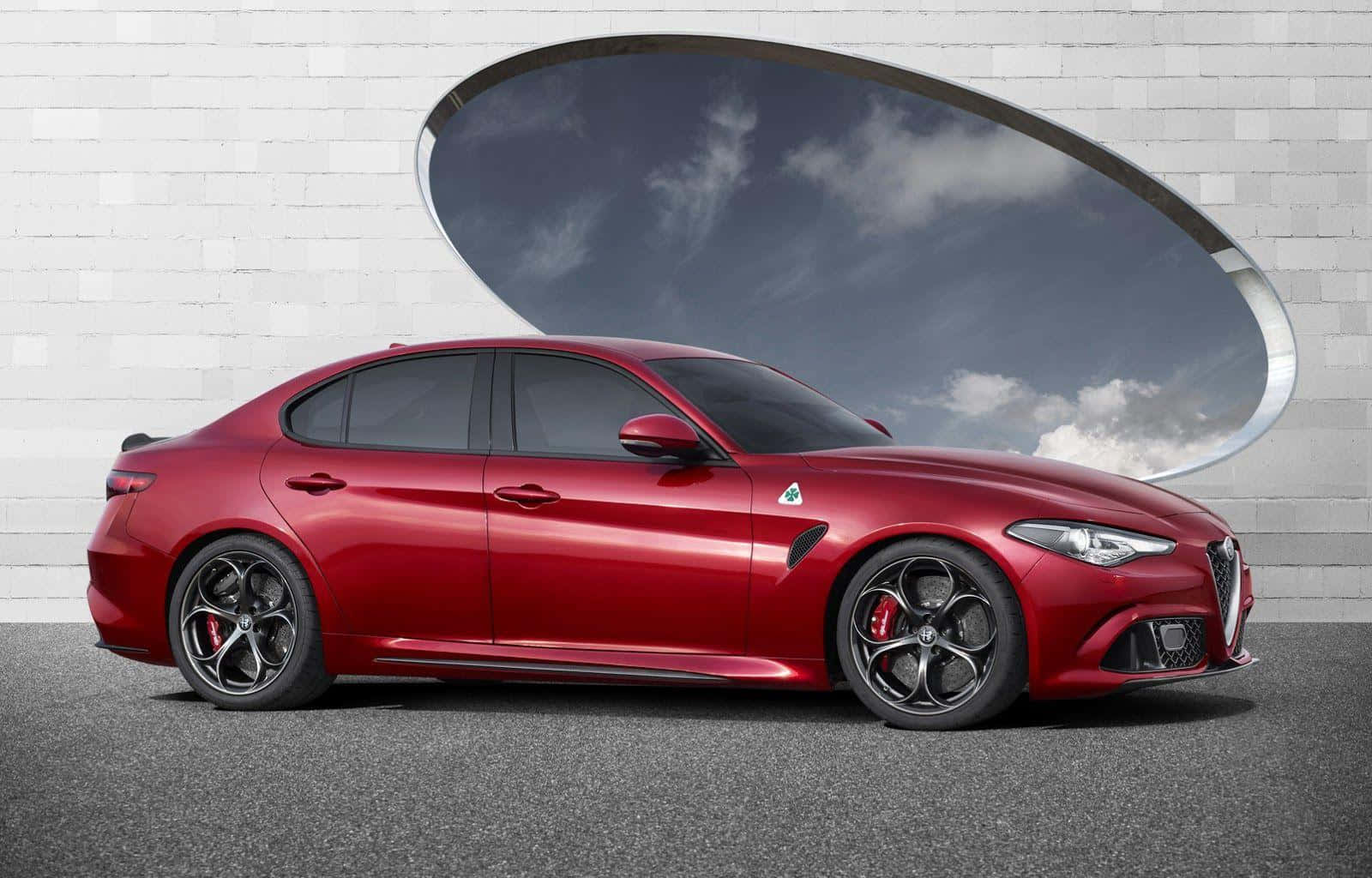 與經典Alfa Romeo探索駕駛的刺激 壁紙 — 一架光澤的紅色 Alfa Romeo 房車停泊在白色磚牆前，牆上有一個大橢圓形窗戶，框著多雲的天空。