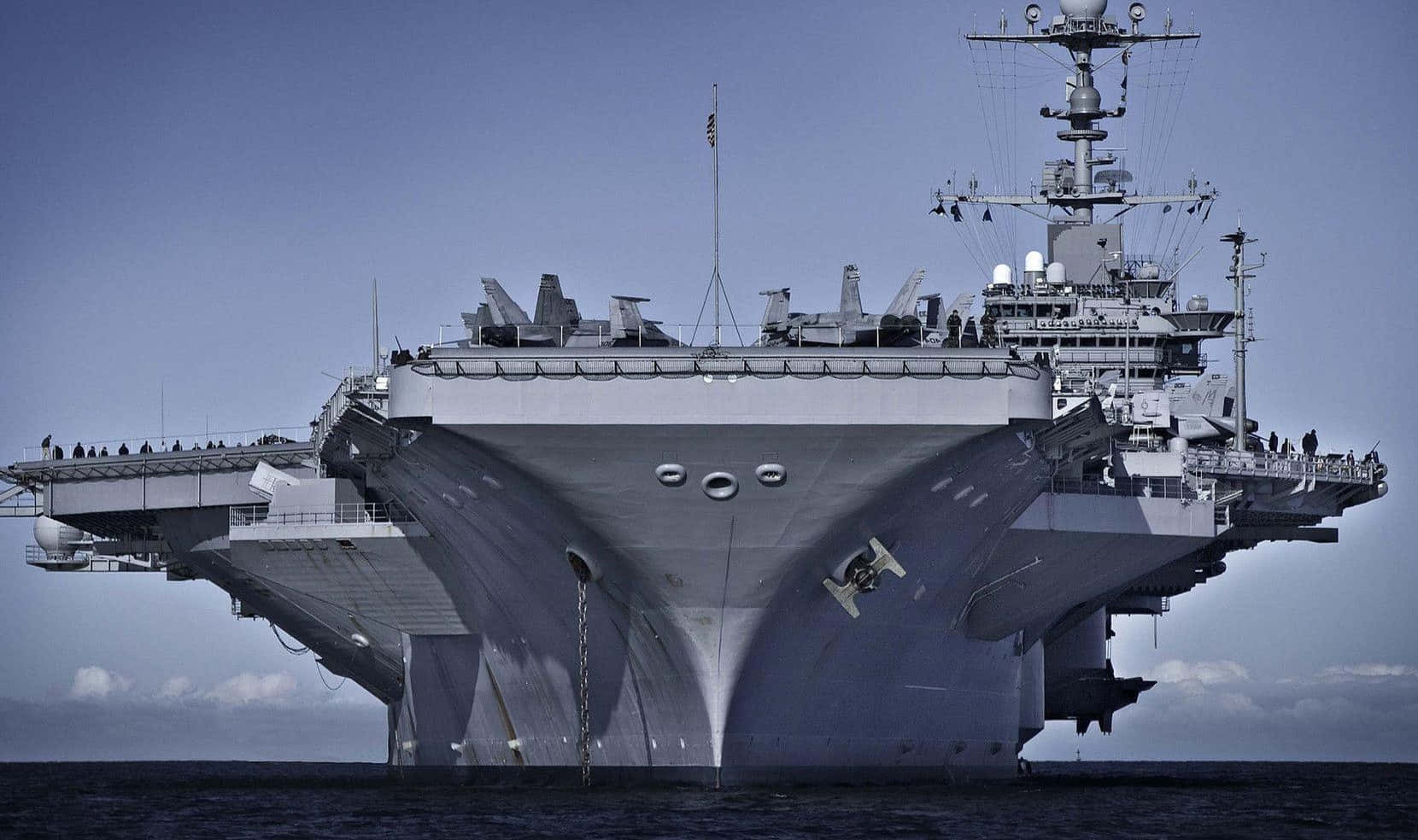 與美國海軍一同探索海洋與更遠方 壁紙 — 一架巨大的灰色航空母艦佔據畫面，其傾斜的飛行甲板載有戰鬥機，背景是清澈的藍天。