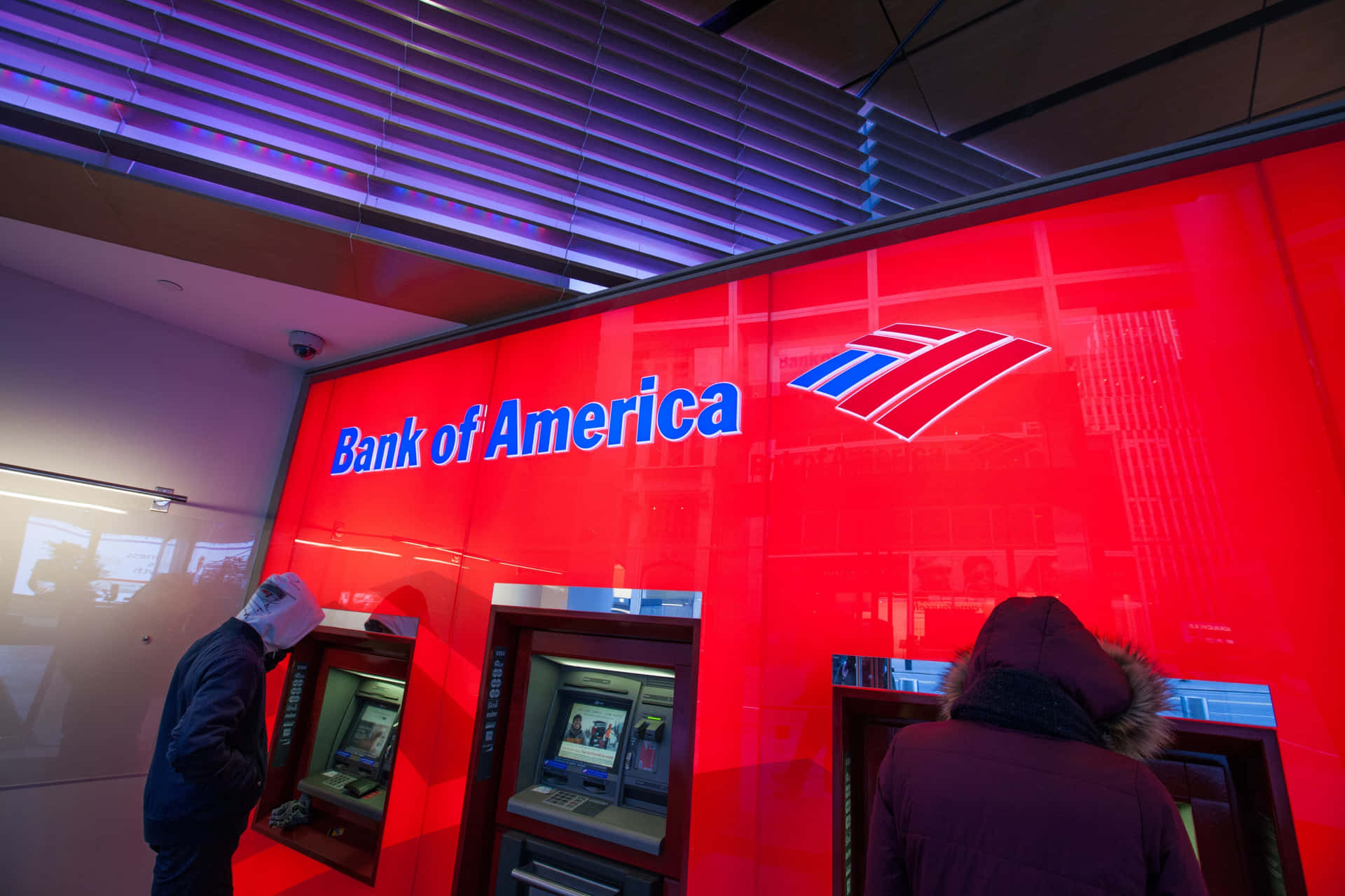 兩個喺紅色牆壁同紫色天花板燈光嘅Bank of America分行內使用自動櫃員機嘅人。