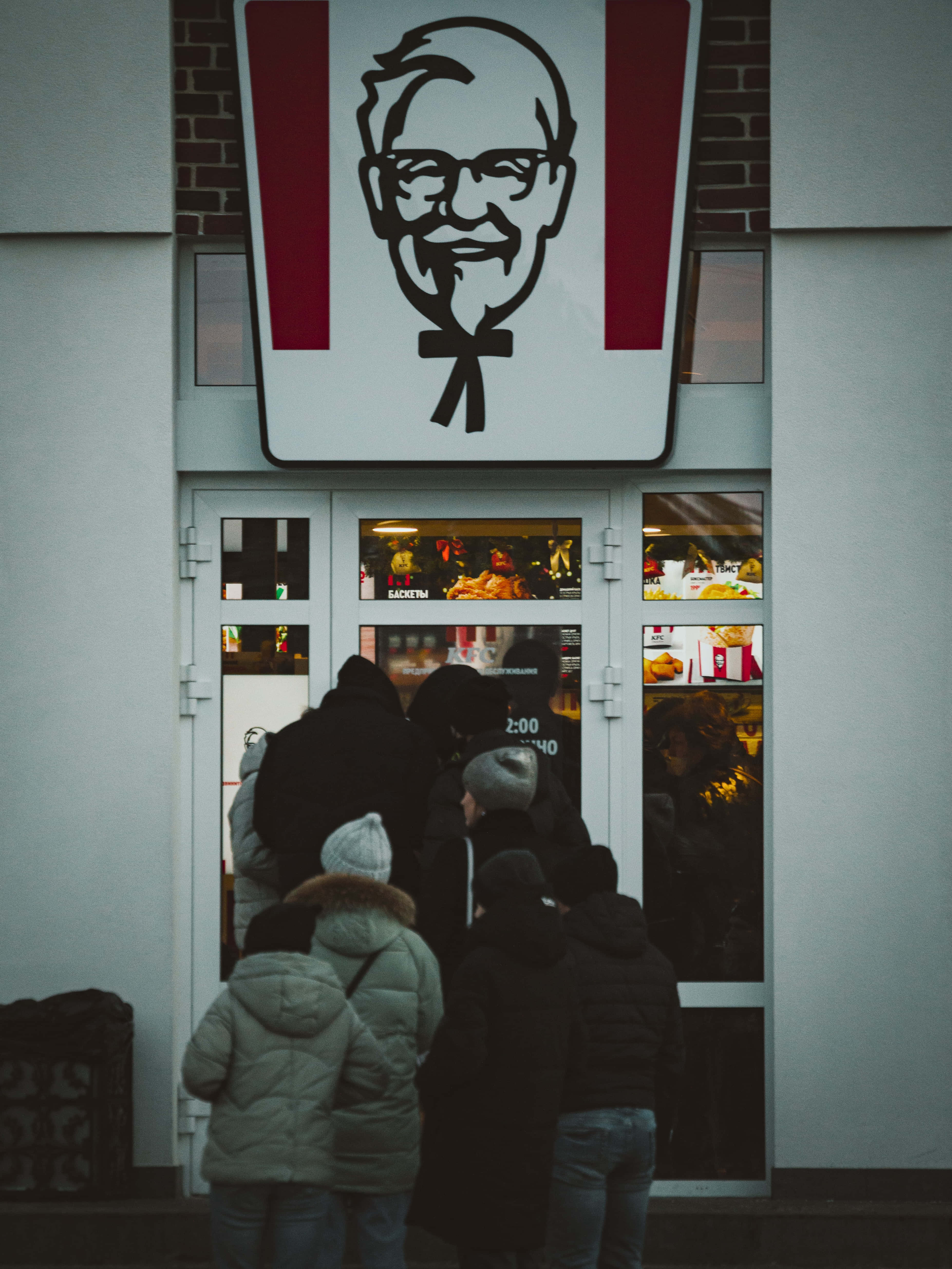與肯德基一起享受美味可口的餐點 壁紙 — 一群穿著冬季外套的人喺一個大型山德士上校招牌下嘅KFC餐廳外面等候。