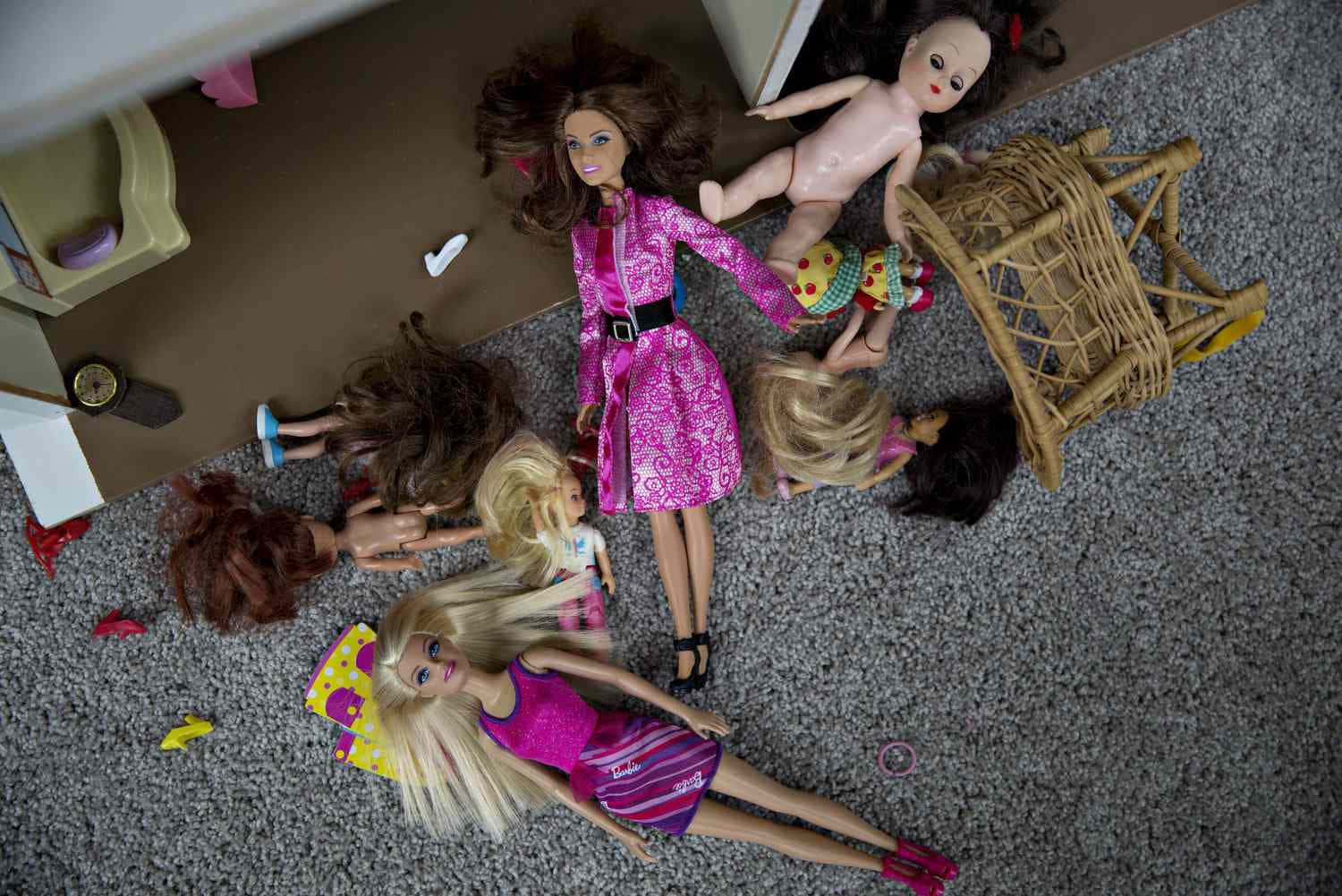 與芭比一起探索世界 壁紙 — Overhead view of scattered dolls in colorful outfits lying on a gray carpet near a toppled wicker chair.