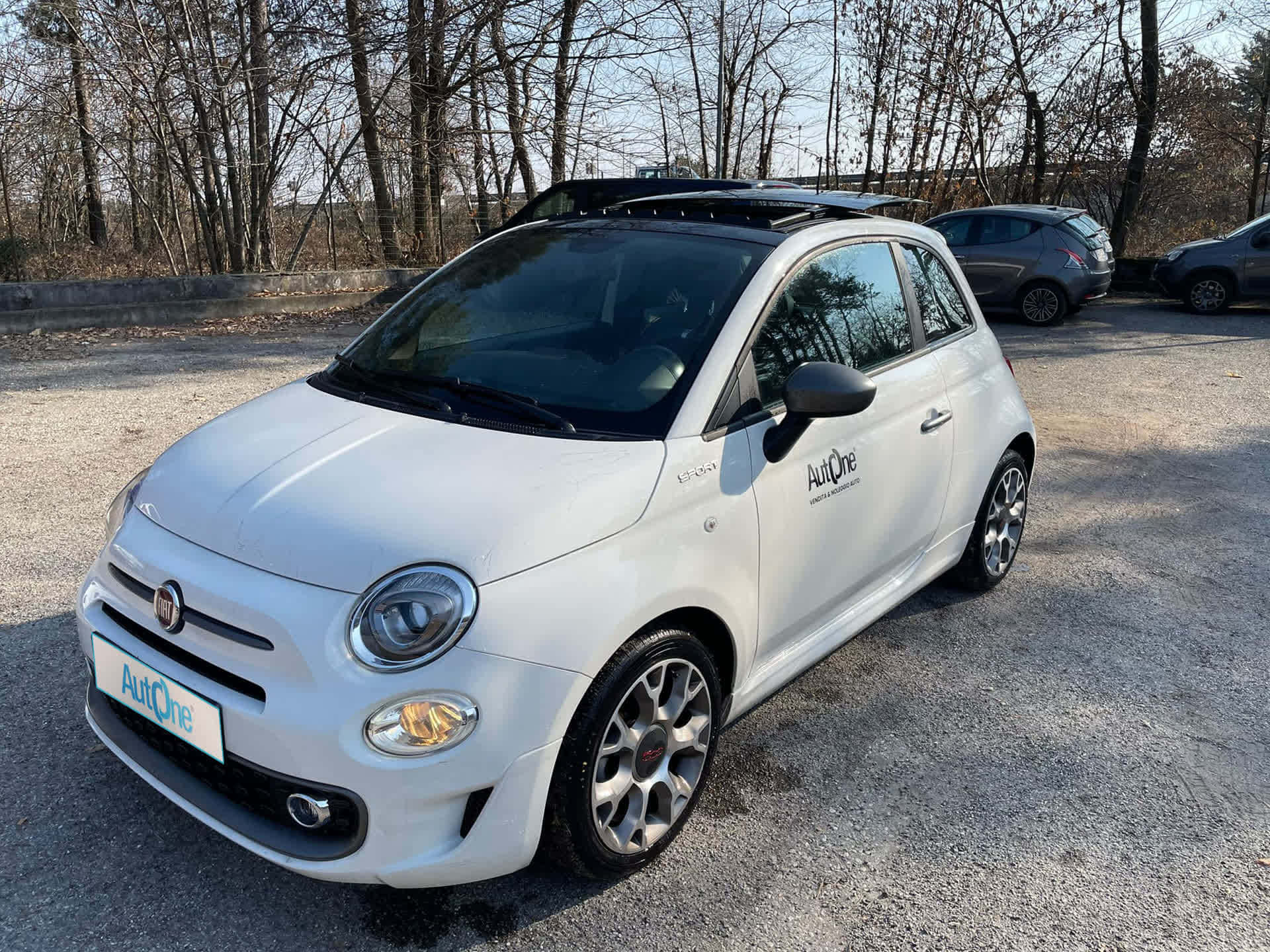 與菲亞特暢遊開闊道路 壁紙 — 一輛白色Fiat 500停在碎石上，背景有光禿禿的樹木，在晴朗的日光下。