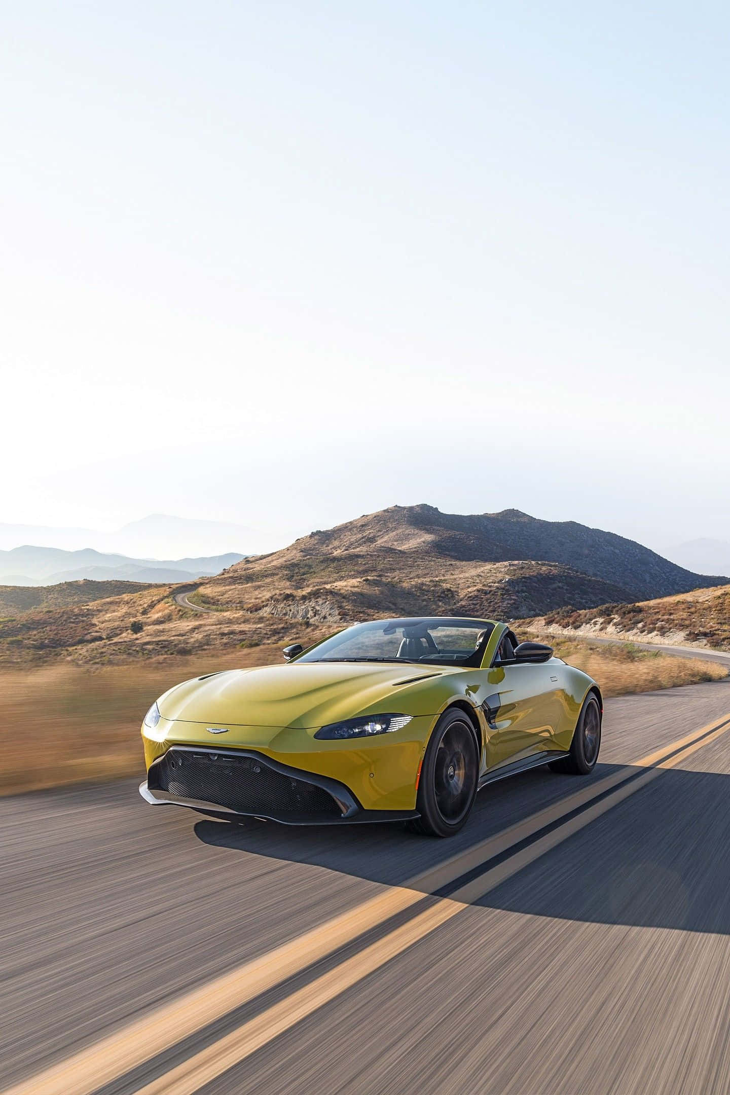與著名阿斯頓·馬丁一同體驗奢華 壁紙 — 一輛鮮黃色的Aston Martin Vantage開篷跑車在晴朗的天空下沿著蜿蜒的山路飛馳。