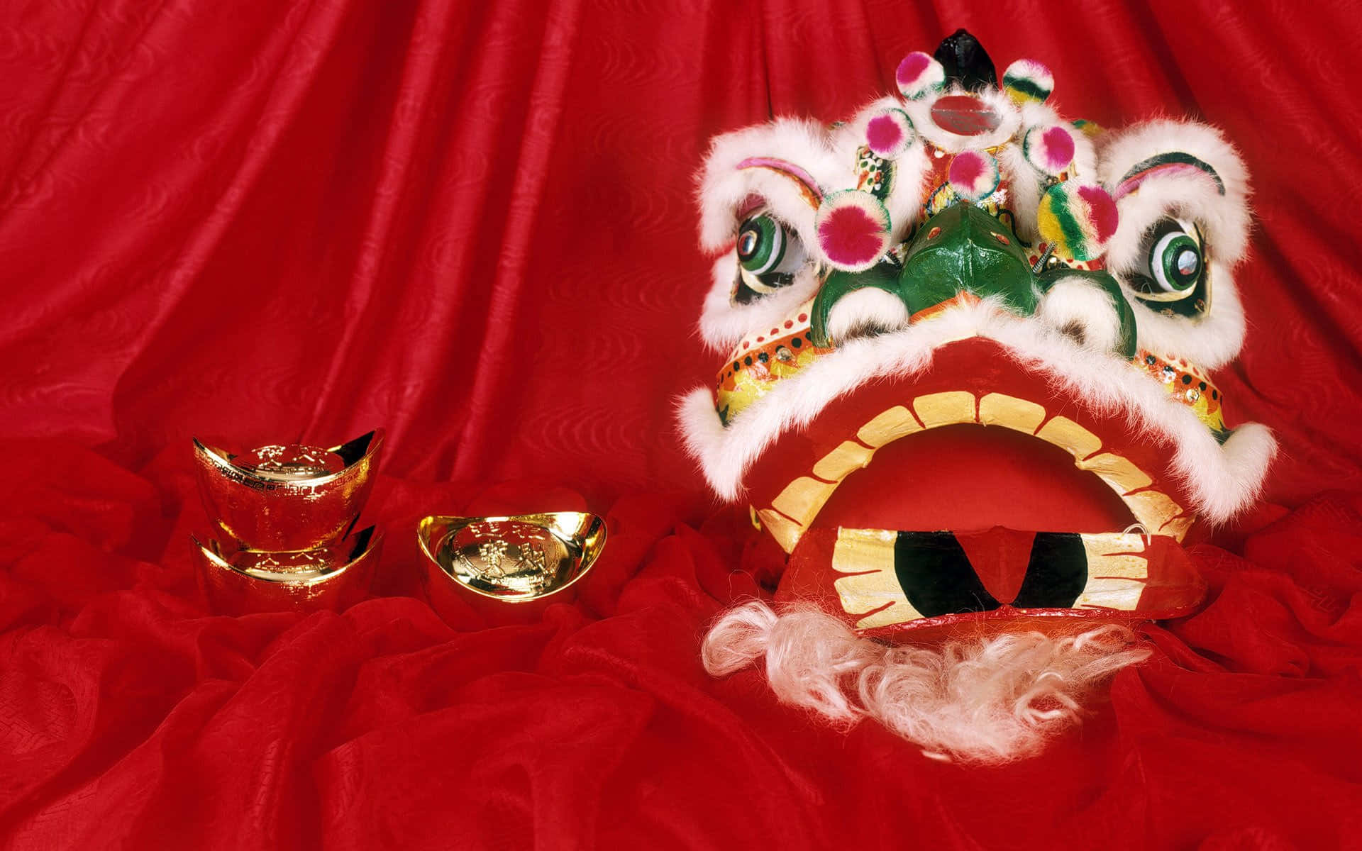 與親友共慶農曆新年 壁紙 — 一個色彩繽紛、眼睛鮮豔的中國舞獅頭，搭配金元寶，放置在濃郁的紅色天鵝絨背景上。