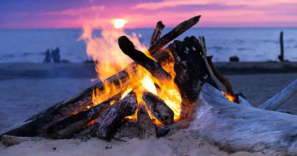 與親友圍爐取暖，享受溫暖時光 壁紙 — 沙灘上，營火在日落時分熊熊燃燒，在紫色和橙色的天空映襯下散發出溫暖的光芒。