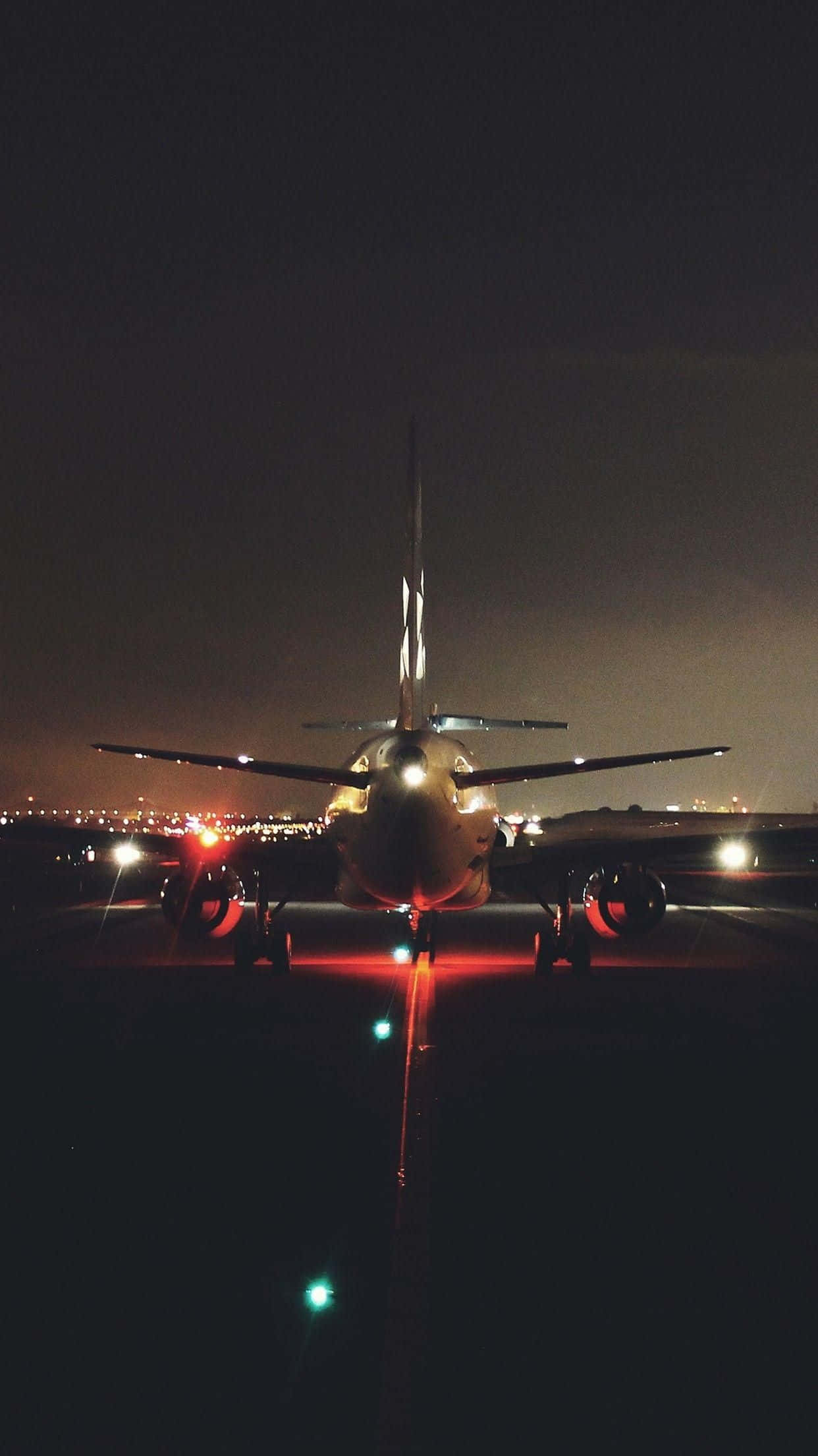 與飛機一同探索天空 壁紙 — 一架商用飛機在夜間停在跑道上，背景有發光的紅燈和遠處的城市燈光。