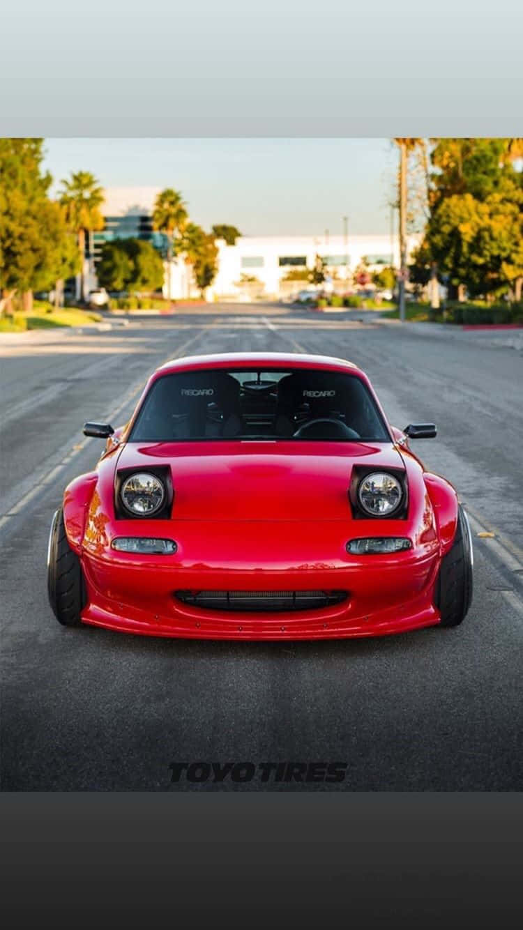 與馬自達MX-5 Miata享受驚艷駕駛 壁紙 — 一輛鮮紅色改裝跑車配備寬輪胎，停放在陽光明媚、兩旁種滿棕櫚樹的街道中央。