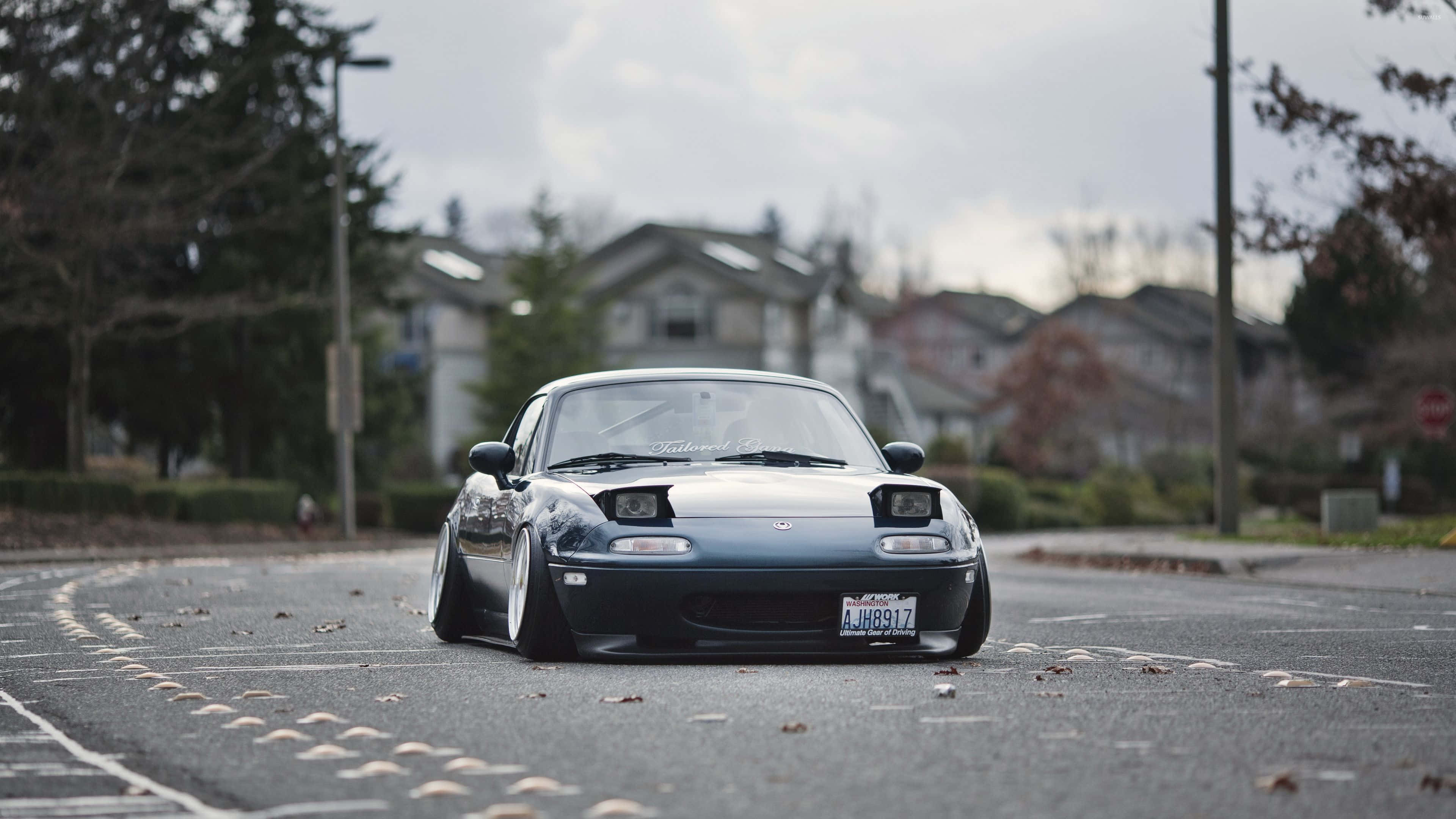 與馬自達MX-5 Miata享受敞篷駕駛樂趣 壁紙 — 一架時尚的深藍色Mazda Miata跑車，配備降低的懸掛系統，停泊在潮濕的郊區街道中央，街道兩旁種滿樹木和房屋，天空多雲。