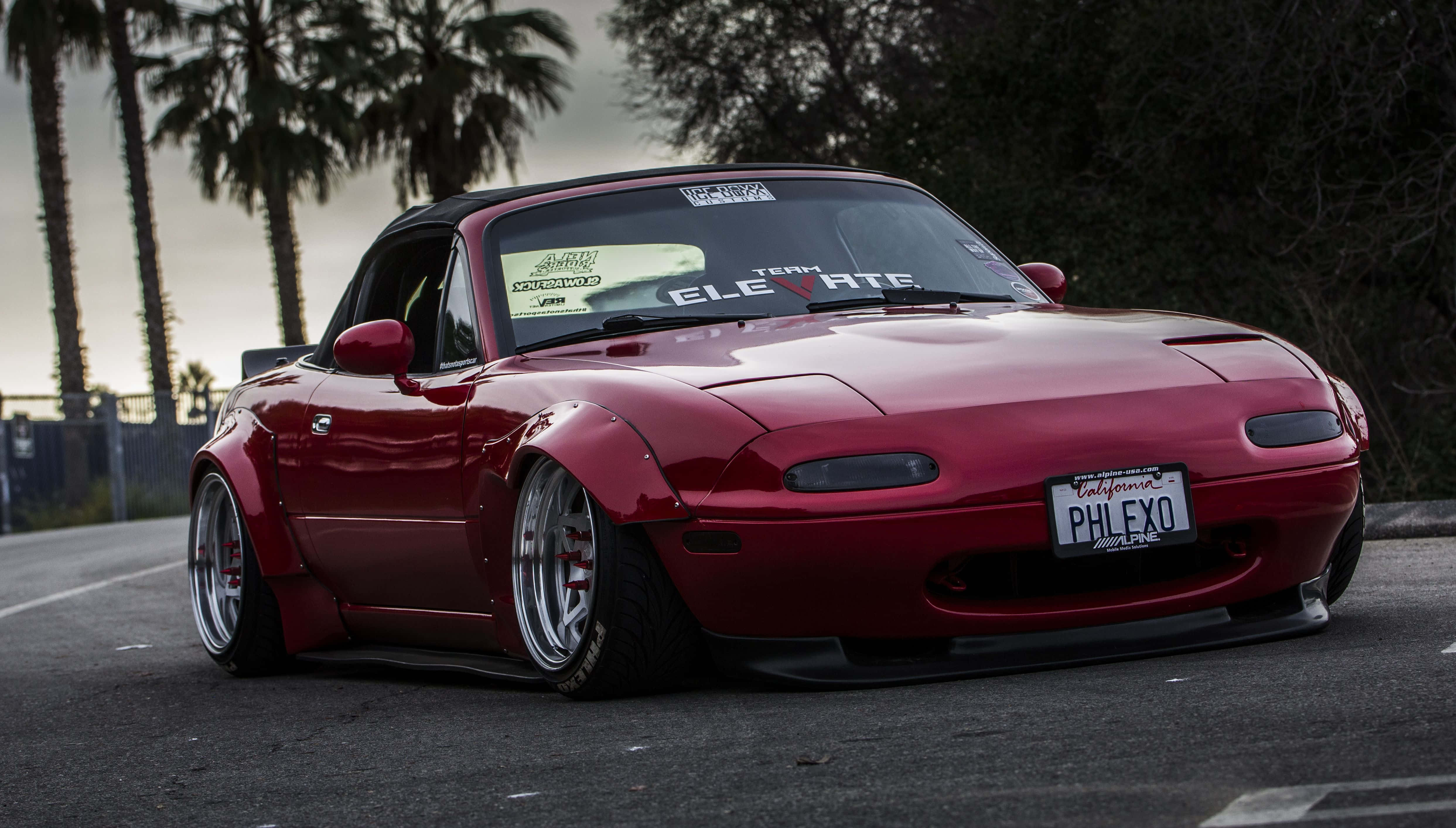 與馬自達MX-5 Miata駛向開放道路 壁紙 — 一架亮紅色改裝萬事得MX-5開篷車停在瀝青路上，背景有棕櫚樹和多雲的天空。