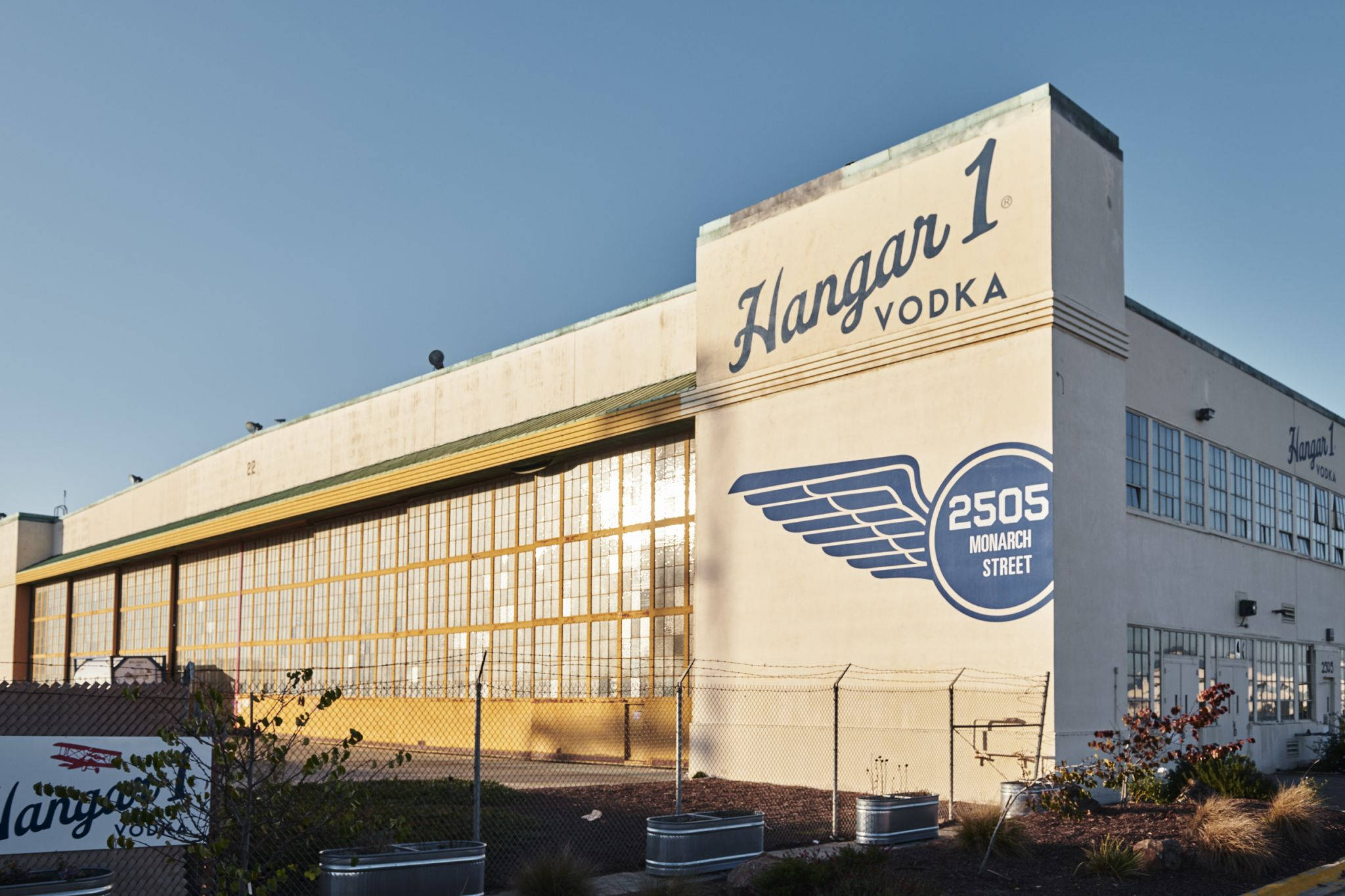 舊式飛機庫中的 Hangar 1 蒸餾廠 壁紙 — 一座大型米色工業建築，帶有 'Hangar 1 Vodka' 品牌和翅膀標誌，在晴朗的藍天下。