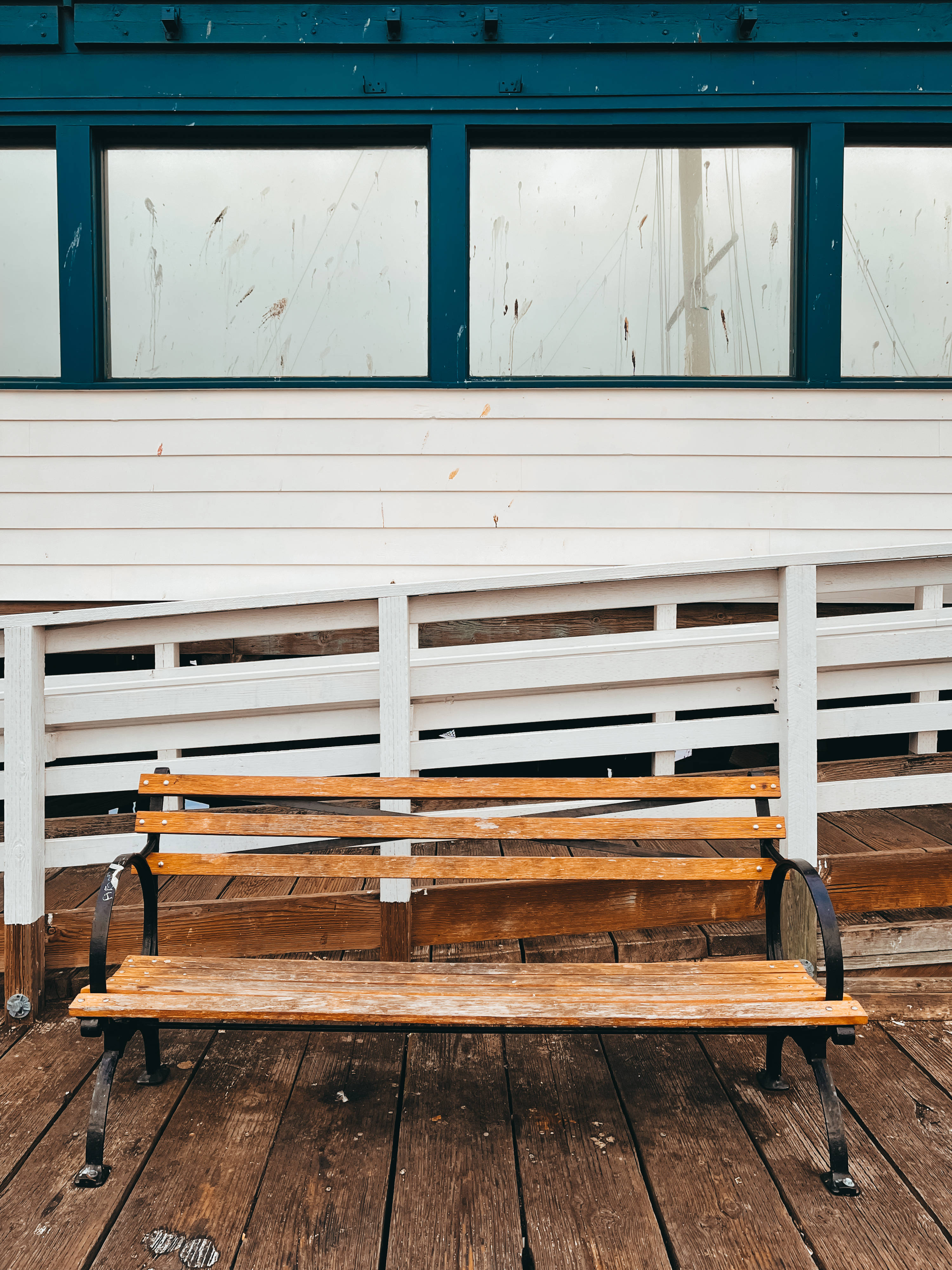 舊火車站木製長椅 iPhone 壁紙 — 一張風化的木製長椅，帶有黑色金屬扶手，放置在深色木甲板上，下方是白色欄杆和青綠色邊框的窗戶。