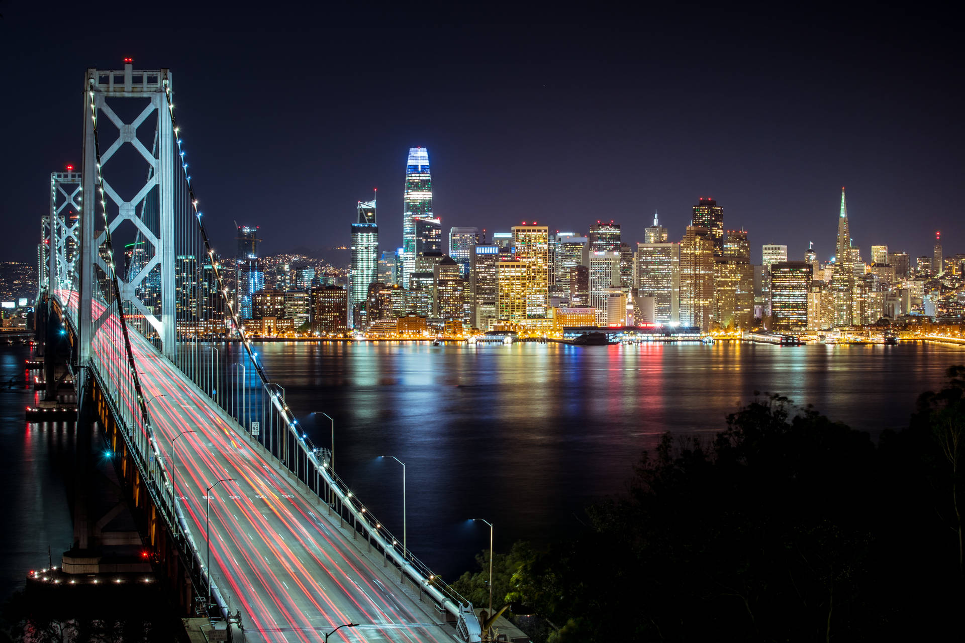 夜間景色，一座亮燈的橋樑在水面上留下燈光軌跡，面向發光的城市天際線。