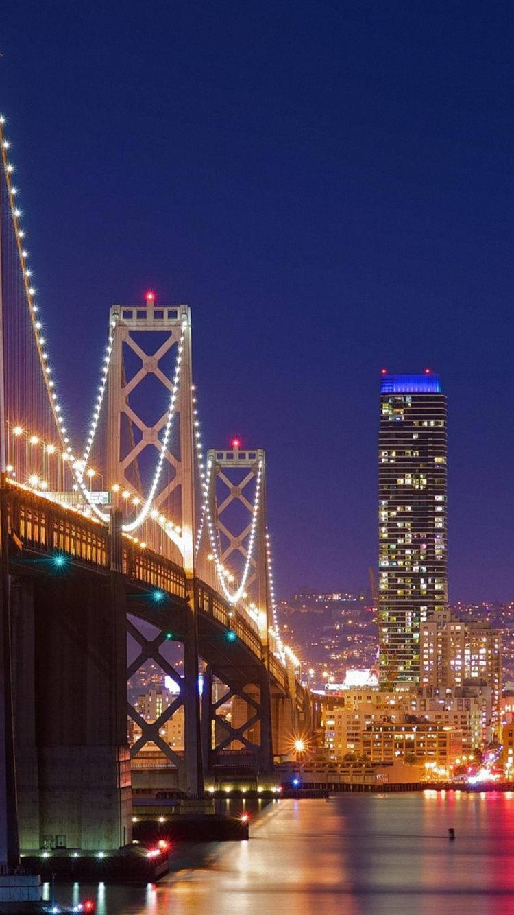 夜間城市景觀，包含一座亮燈的橋樑和摩天大樓，帶有水面的倒影。