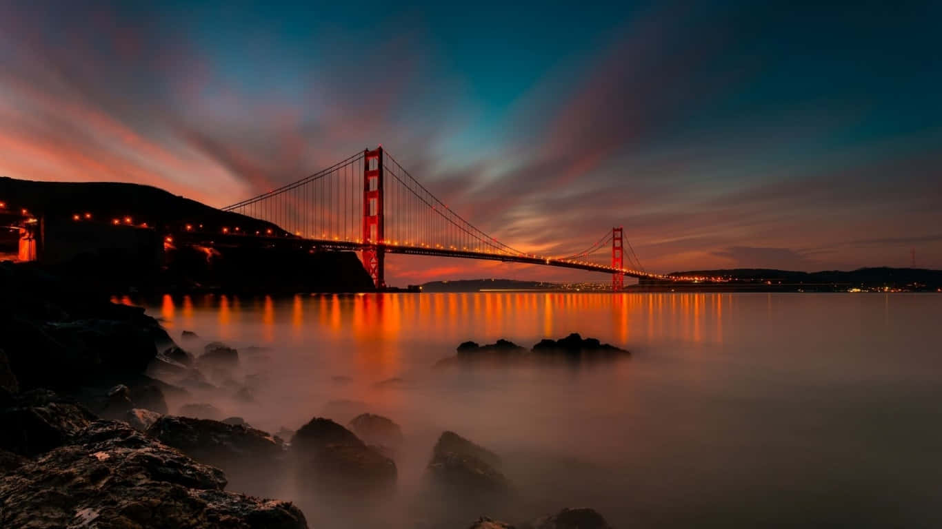 日落時分的金門大橋，燈光在平靜的水面上閃爍倒映，前景有深色岩石。
