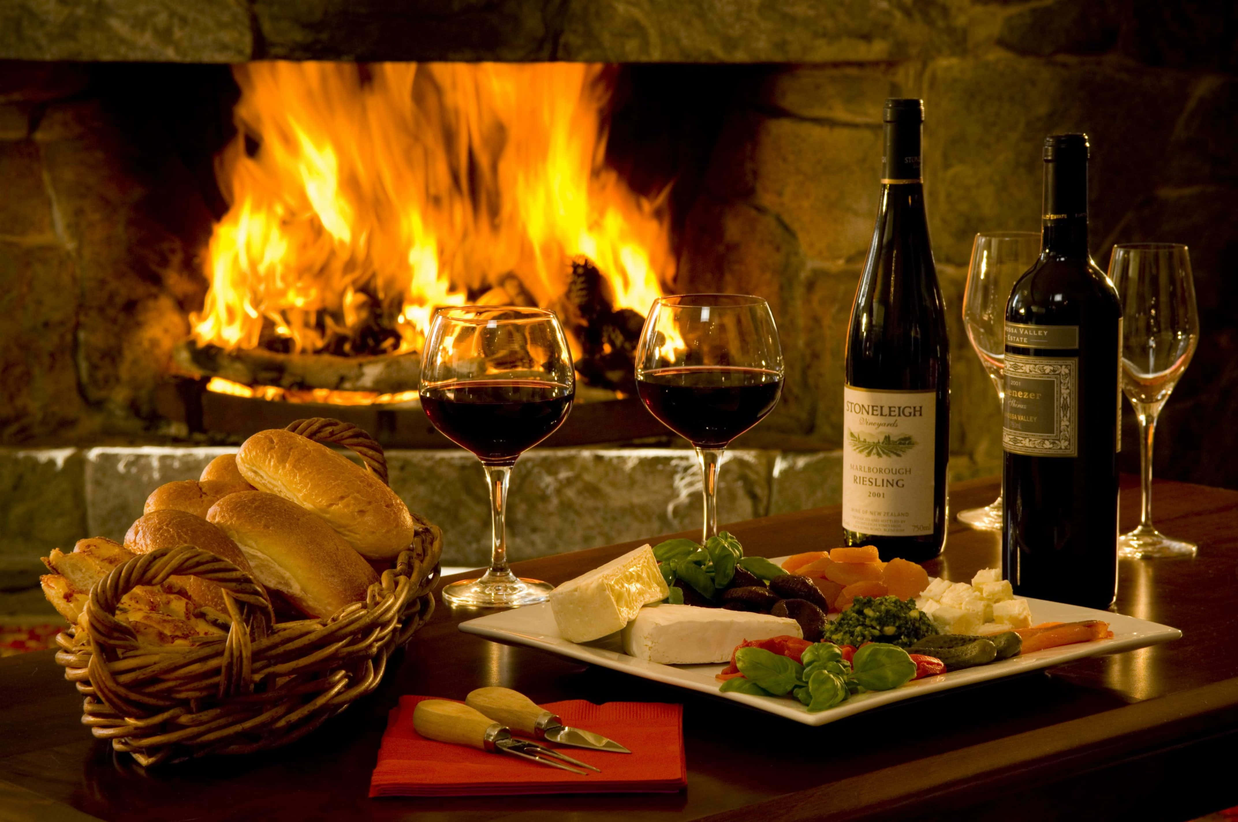 Autumn Fireplace 桌布 壁紙 — 舒適的壁爐光芒照亮了擺有酒瓶、酒杯、芝士、麵包和新鮮香草的木桌。