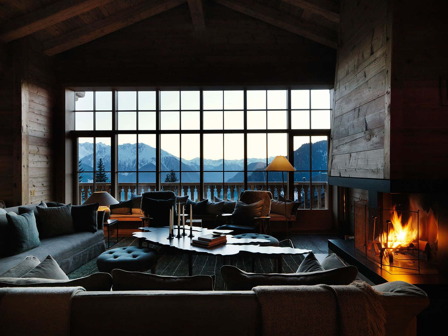 Cozy Fire 桌布 壁紙 — 舒適的山間小屋內部，設有熊熊燃燒的壁爐，透過大窗戶可欣賞白雪皚皚的山峰全景。