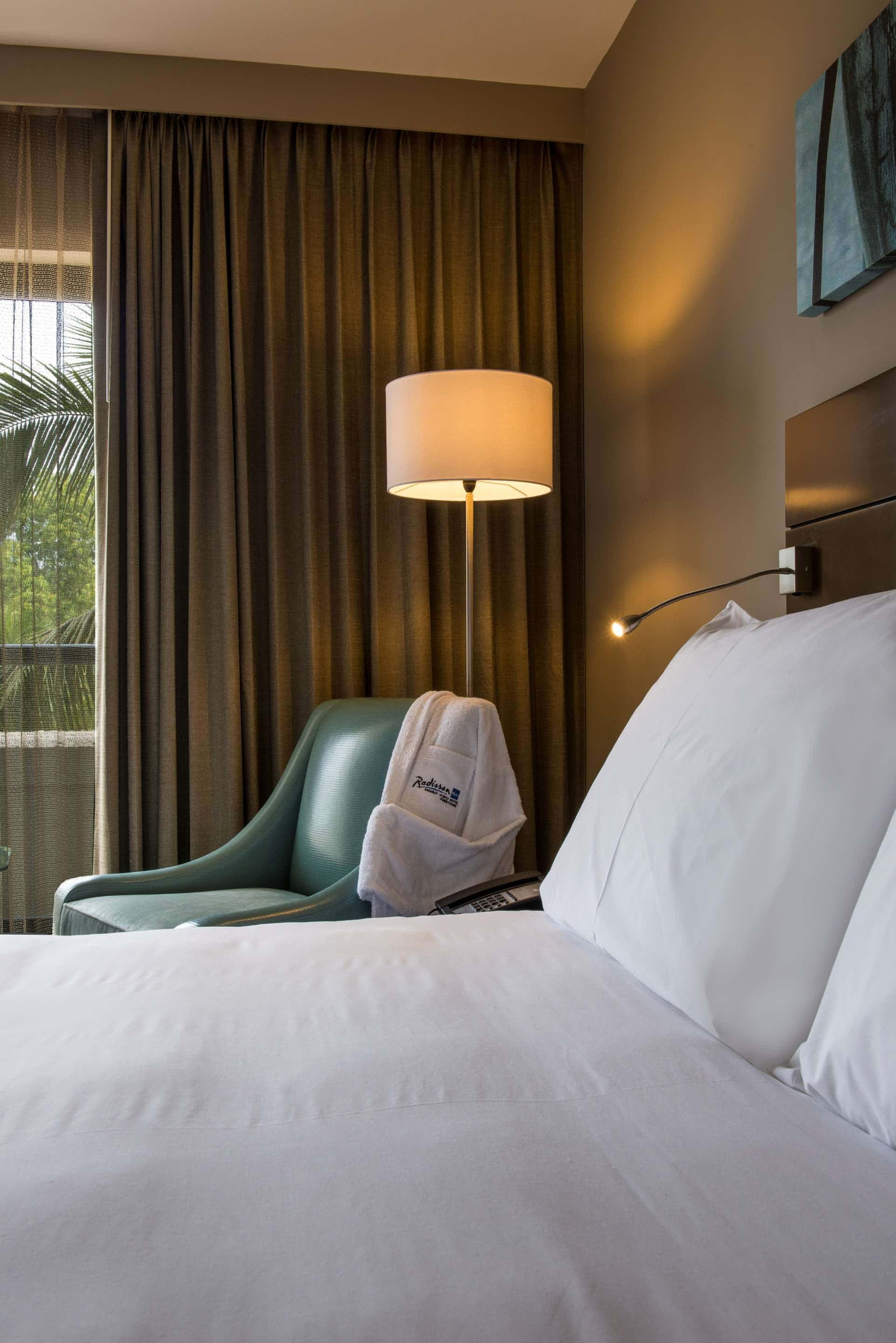 Hotel Room 桌布 壁紙 — 舒適的酒店客房，配有整齊的床鋪、掛著浴袍的藍綠色椅子，以及來自落地燈和壁燈的溫暖燈光。